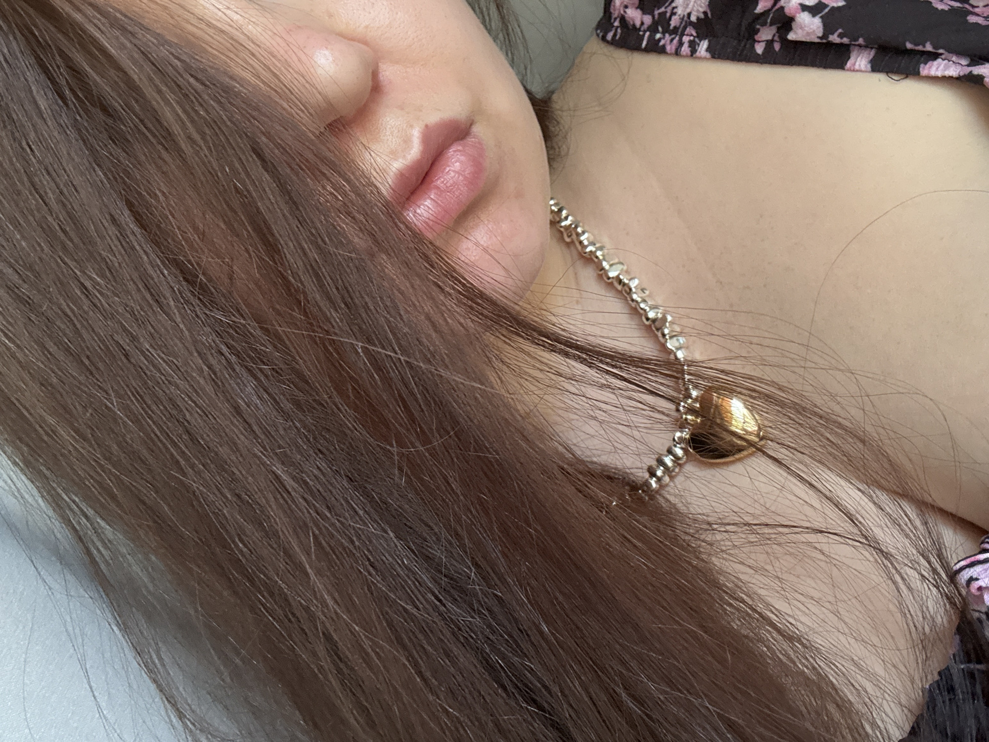 The image is a close-up shot of a person's neck and lower face, with their hair obscuring part of their face. The person is wearing a silver chain necklace with a gold heart-shaped pendant. They are also wearing a dark-colored top with a floral pattern. The background appears to be a plain, light-colored wall. There is no indication of the location being in Chaikovsky, Russia, or any other specific city.