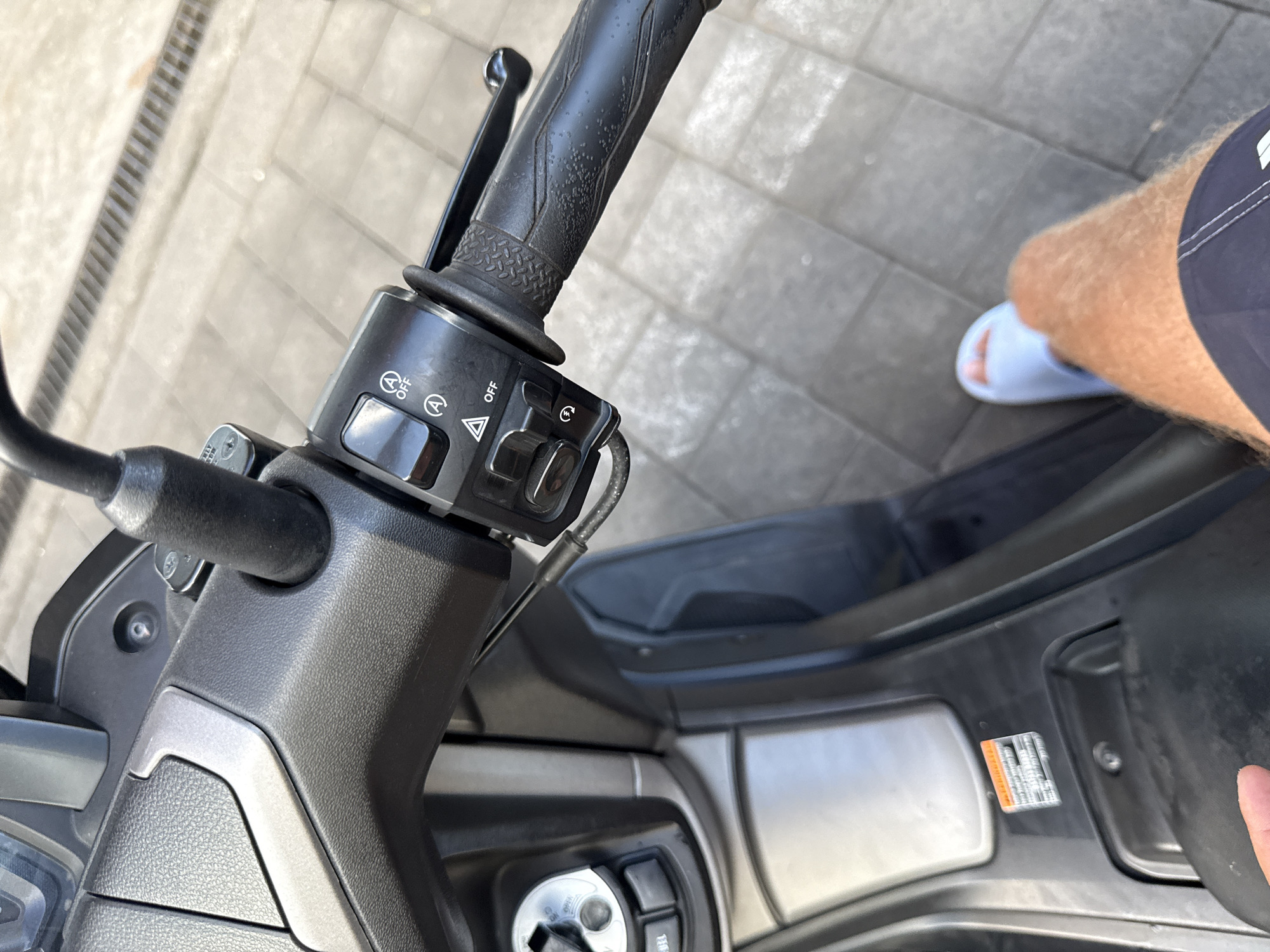 A close-up, high-angle shot captures the handlebars and central console of a parked scooter in Ungasan, Indonesia. The handlebars, with a textured black grip, feature a switch cluster including indicators, headlight settings, and a starter button. The central console includes the ignition and warning stickers. In the periphery, a portion of a person's leg, clad in dark shorts and a white sandal, is visible, suggesting someone is standing beside the scooter. The background consists of a paved surface.