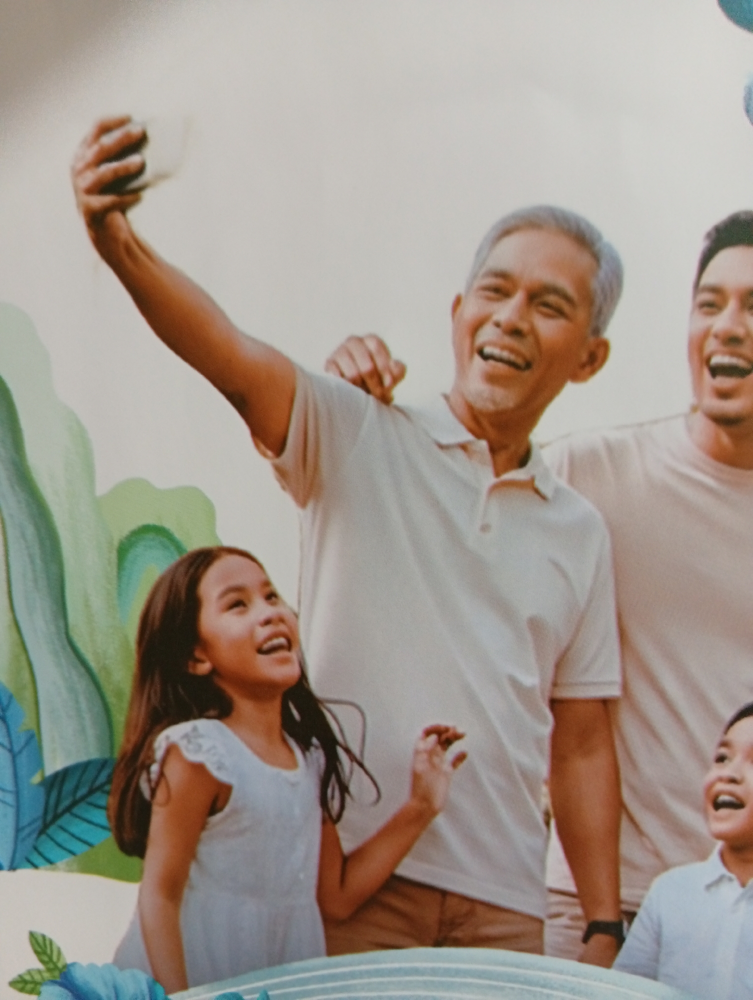 The image shows a candid, eye-level outdoor portrait of a multi-generational family of four, posing for a selfie. A man in his 60s with graying hair and a light-colored polo shirt holds up a smartphone in his right hand, arm extended, to capture the moment. He is smiling broadly, looking towards the camera. To his left, a young girl, around 8 years old, with long dark hair and wearing a white dress, is also smiling and looking upwards, possibly at the phone or her grandfather. Her left arm is raised, with her hand in a gesture of excitement. To the right of the older man, partially obscured, is a younger man, likely his son, in a light-colored t-shirt, also smiling and looking towards the camera. In the lower right corner, another young boy, possibly a second son or nephew, with dark hair and wearing a light blue collared shirt, is also visible and smiling. The background is a soft, out-of-focus blend of light colors, suggesting an outdoor setting with a bright sky. In the mid-ground and left, there are stylized, abstract green and blue leaf-like shapes and blue flowers, implying a decorative or artistic element in the environment. The overall impression is one of joy and familial connection. No specific landmarks within Tlogomulyo, Indonesia are identifiable in the image.