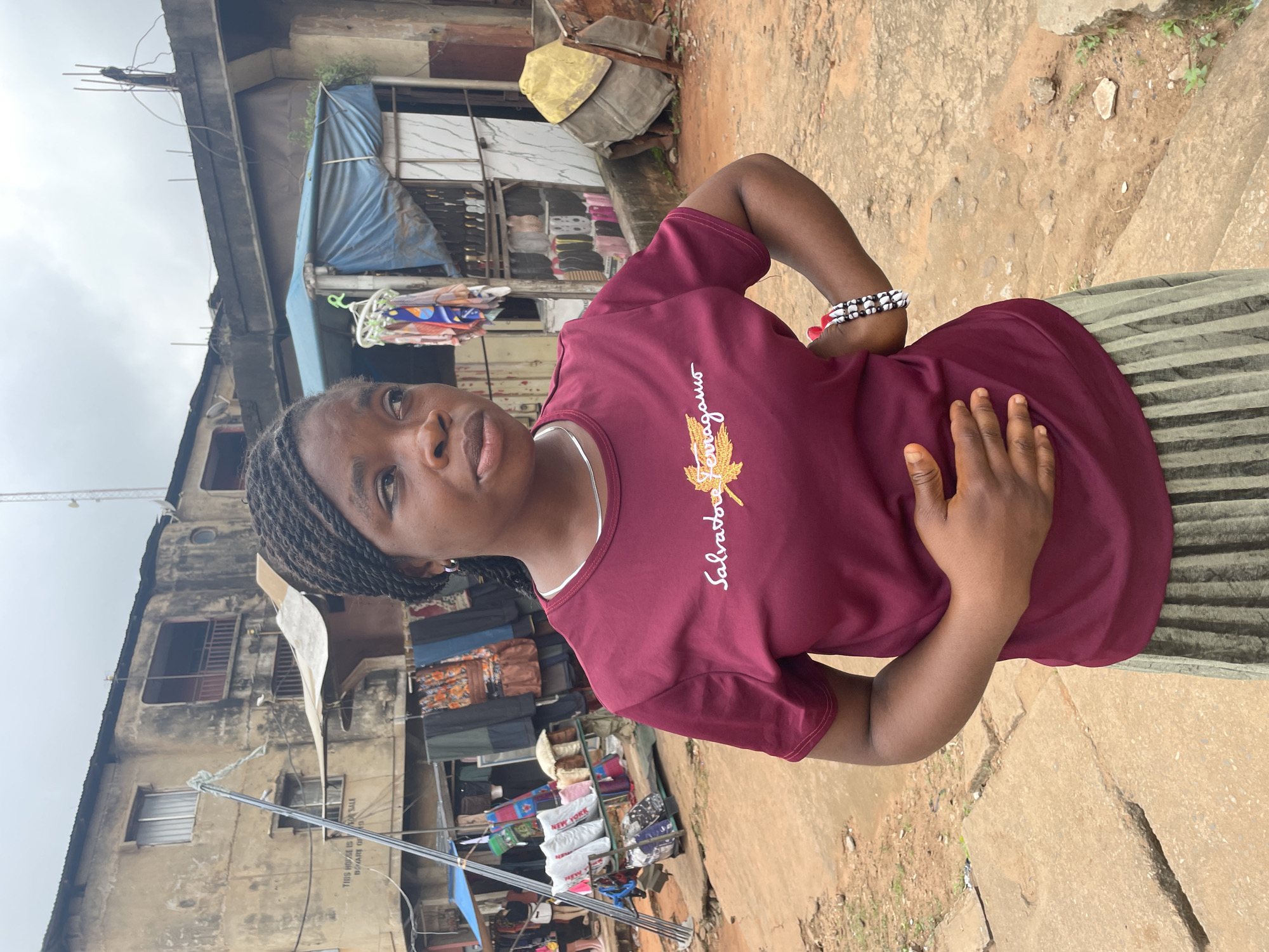 A young Black woman with braided hair stands outdoors in what appears to be a market area. She is wearing a maroon t-shirt with a gold embroidered logo and a green and gray striped skirt. She has her left hand on her stomach and her right hand on her hip. She is wearing a silver necklace and a beaded bracelet on her right wrist. The background shows a weathered, multi-story building with balconies and laundry hanging from lines. In front of the building, there are market stalls with clothing and other items displayed. The ground is a mix of dirt and concrete. The sky is overcast. The image was taken in Nkpor, Nigeria.