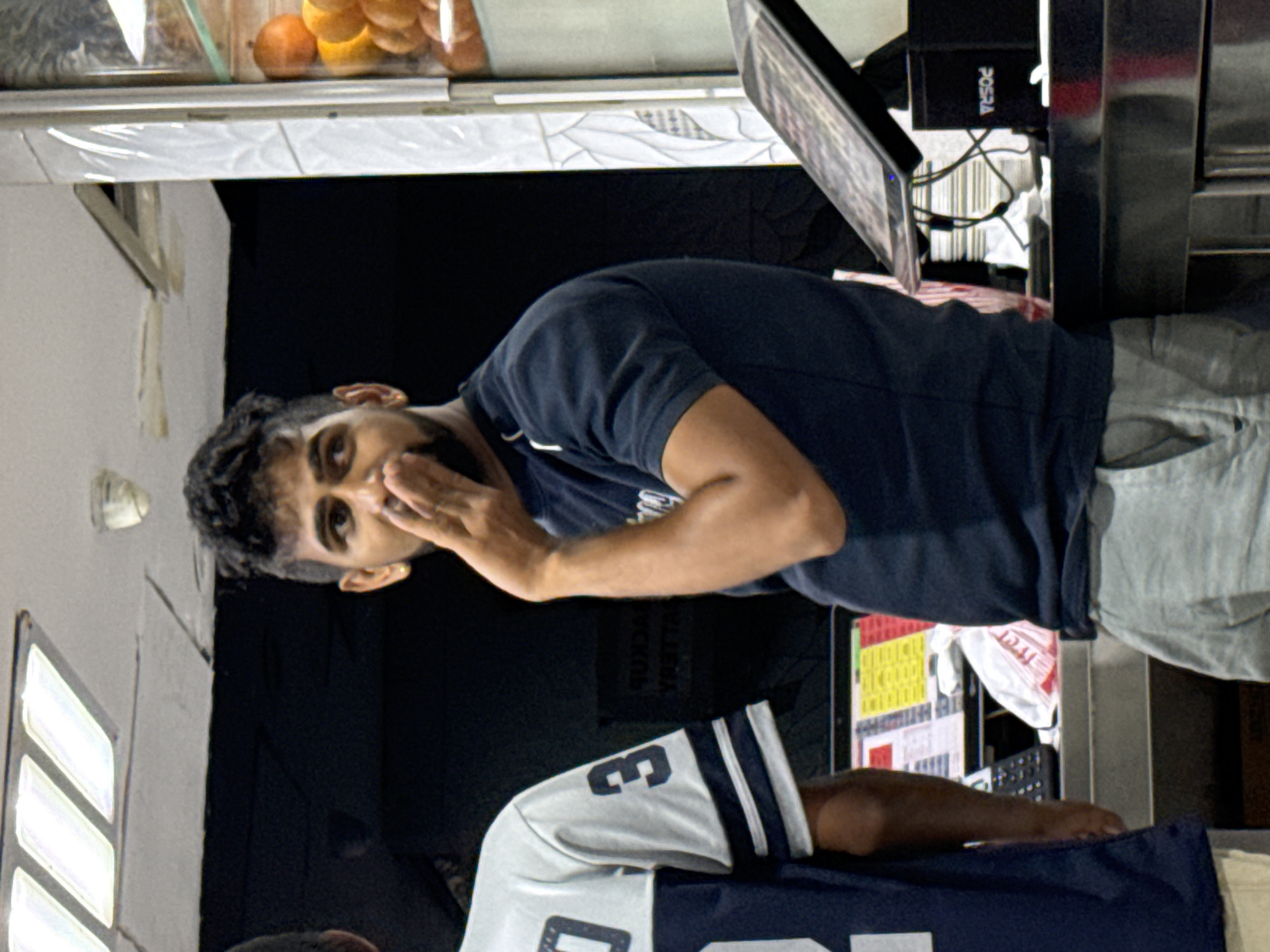 A medium shot captures two men standing behind a counter in what appears to be a food establishment. The man on the right, in focus, is wearing a dark blue t-shirt and light grey pants. He has dark, curly hair and a beard, and is touching his mouth with his right hand, looking to his left with a curious expression. A gold earring is visible in his left ear. The man on the left is wearing a white and blue jersey with the number '3' on the sleeve. His face is not visible. The background is dark, with fluorescent lighting fixtures visible on the ceiling. A display of oranges and other produce is seen through a glass partition to the right. Various items are on the counter, including a digital display, a black cash register, and a white plastic bag. The overall setting suggests a casual, possibly busy, retail environment in Dubai, United Arab Emirates.