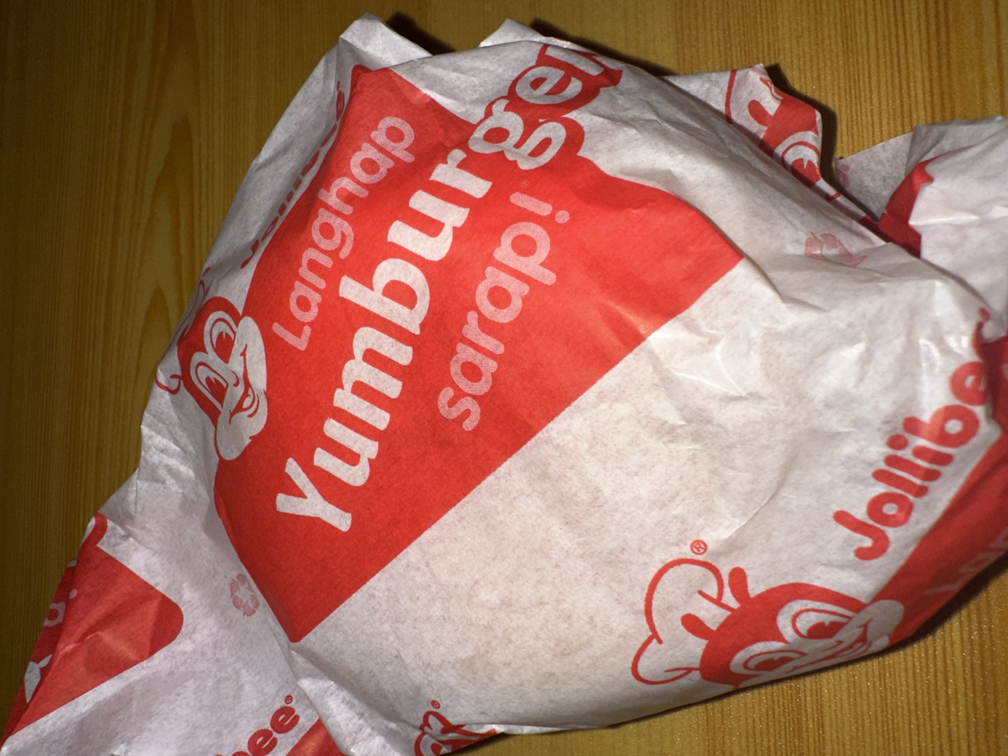 The image shows a Jollibee Yumburger wrapped in its branded white paper wrapper with red printing, including the Jollibee logo and the words "Langhap Yumburger sarap!" It is placed on a wooden surface, possibly a table. The location is likely Davao City, Philippines, given that Jollibee is a popular fast-food chain in the Philippines.