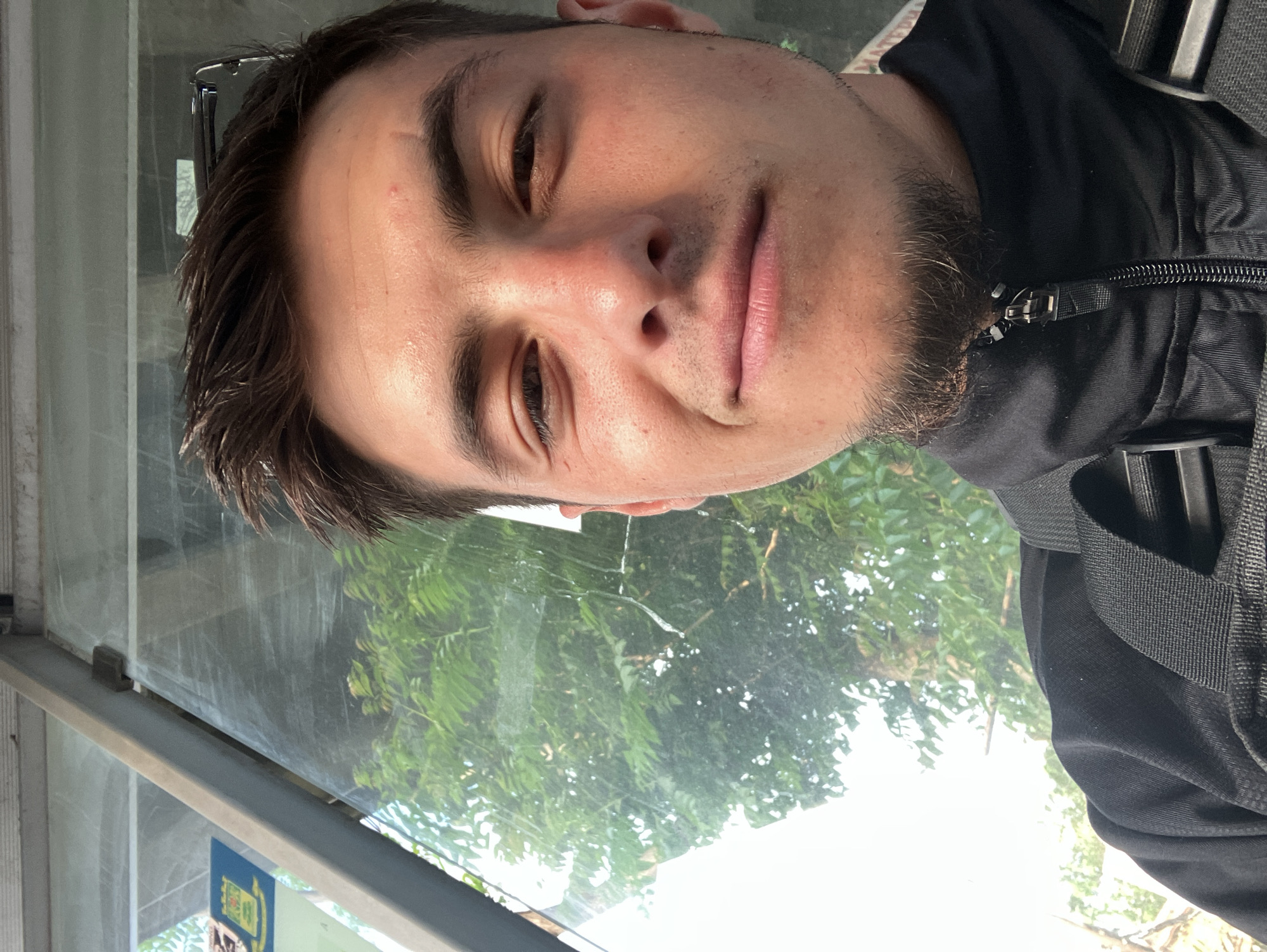 A selfie shows a man with brown hair and a stubbly beard, wearing a black jacket. The background is out of focus, but it appears to be foliage visible through a glass surface, potentially a bus shelter or similar structure. There is a sign with yellow and blue colors. This image was taken in a city, possibly Plovdiv, Bulgaria.