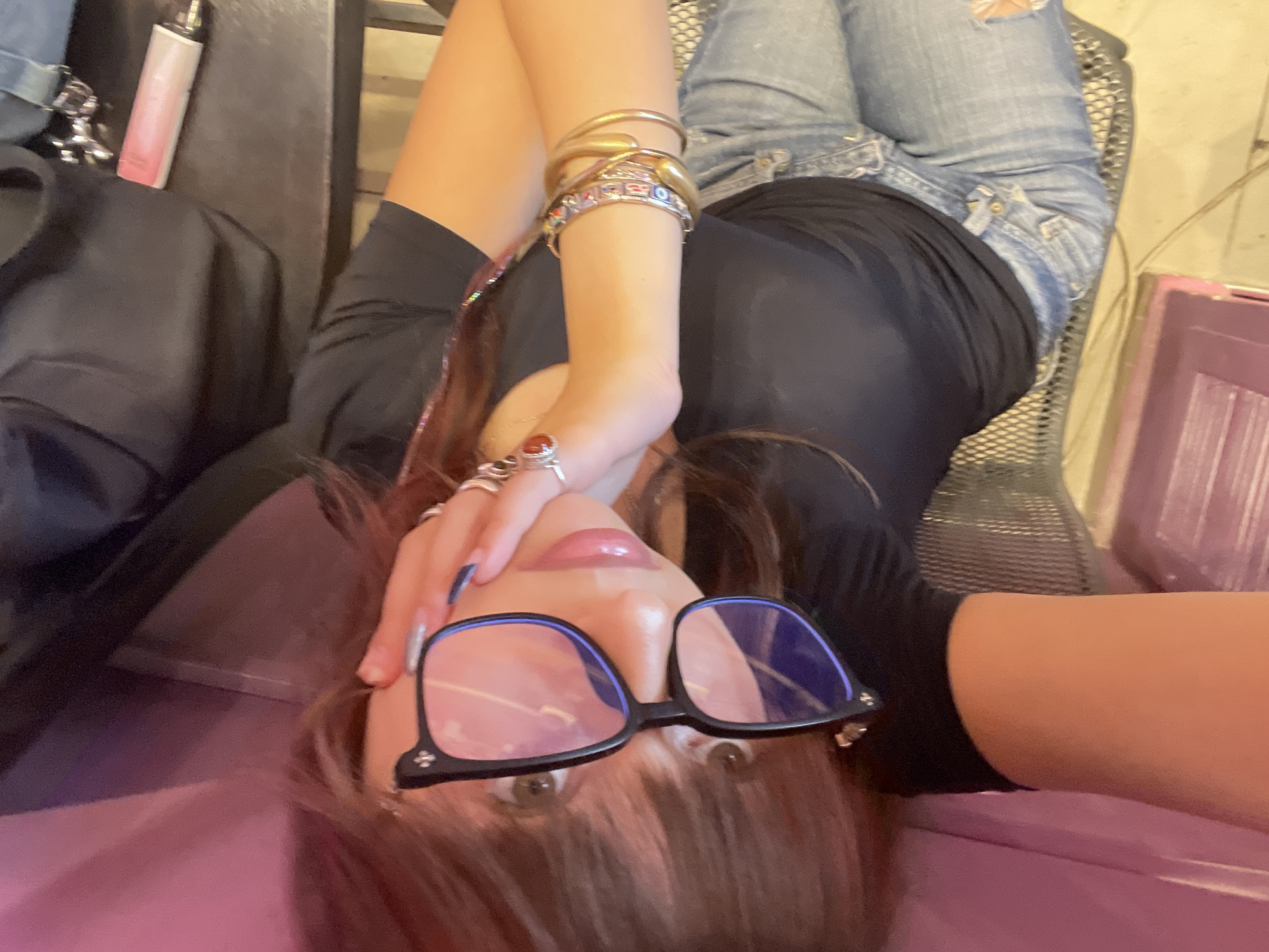 A person is lying down, holding their hand to their face and wearing glasses and jewelry. They are wearing a black t-shirt and blue jeans. The environment appears to be indoors, possibly a cafe or salon, with a pink surface and a black table. The image is taken from a high angle, with the person in the foreground. The person's face is partially obscured by their hand and glasses.