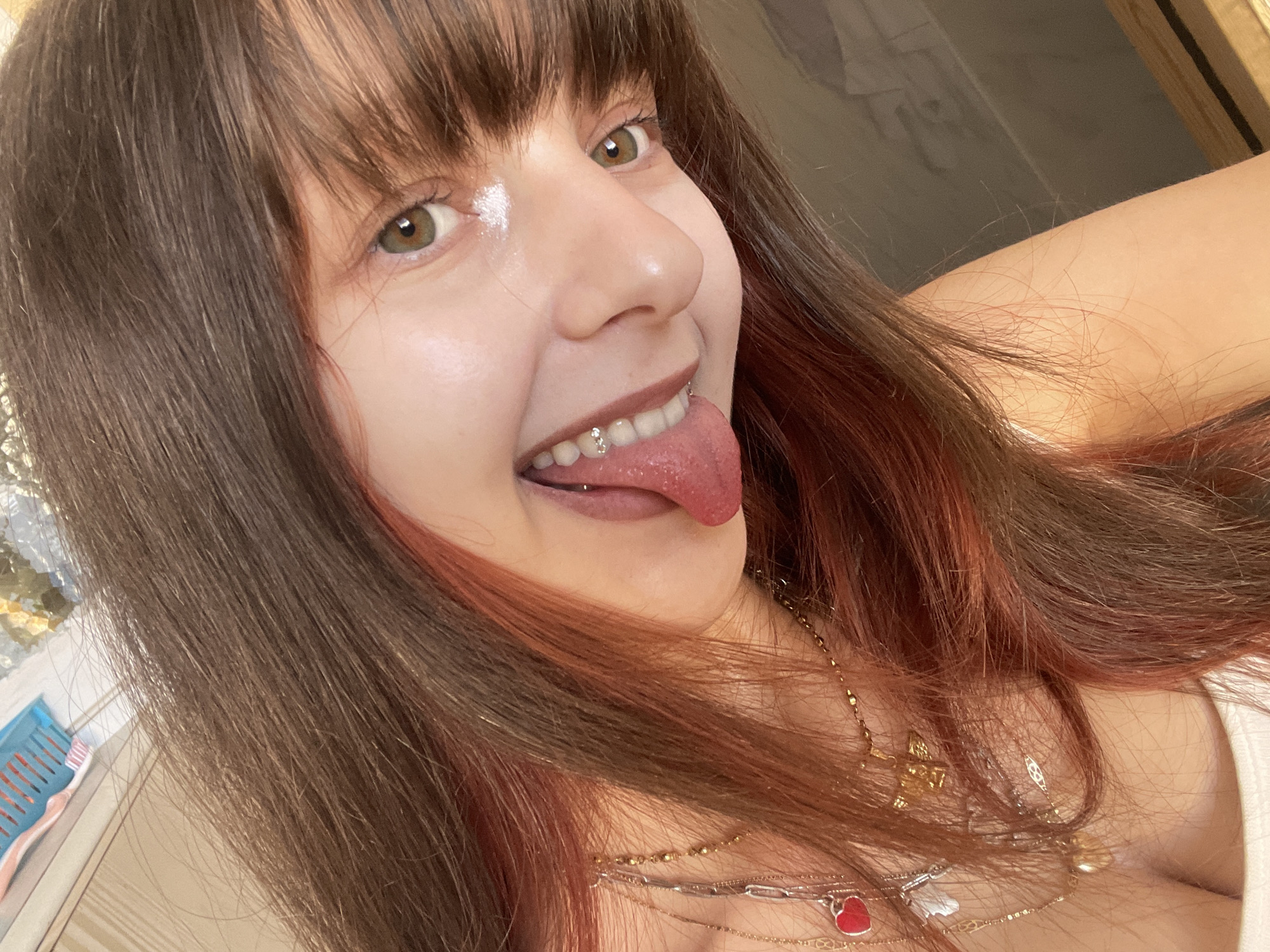 A person with light skin, brown hair with reddish highlights, and green eyes is looking at the camera with their tongue sticking out, revealing a tongue piercing. They are wearing multiple necklaces. The background is indistinct. There is no information available to discern the location in Petah Tikva, Israel, or any other city or location.