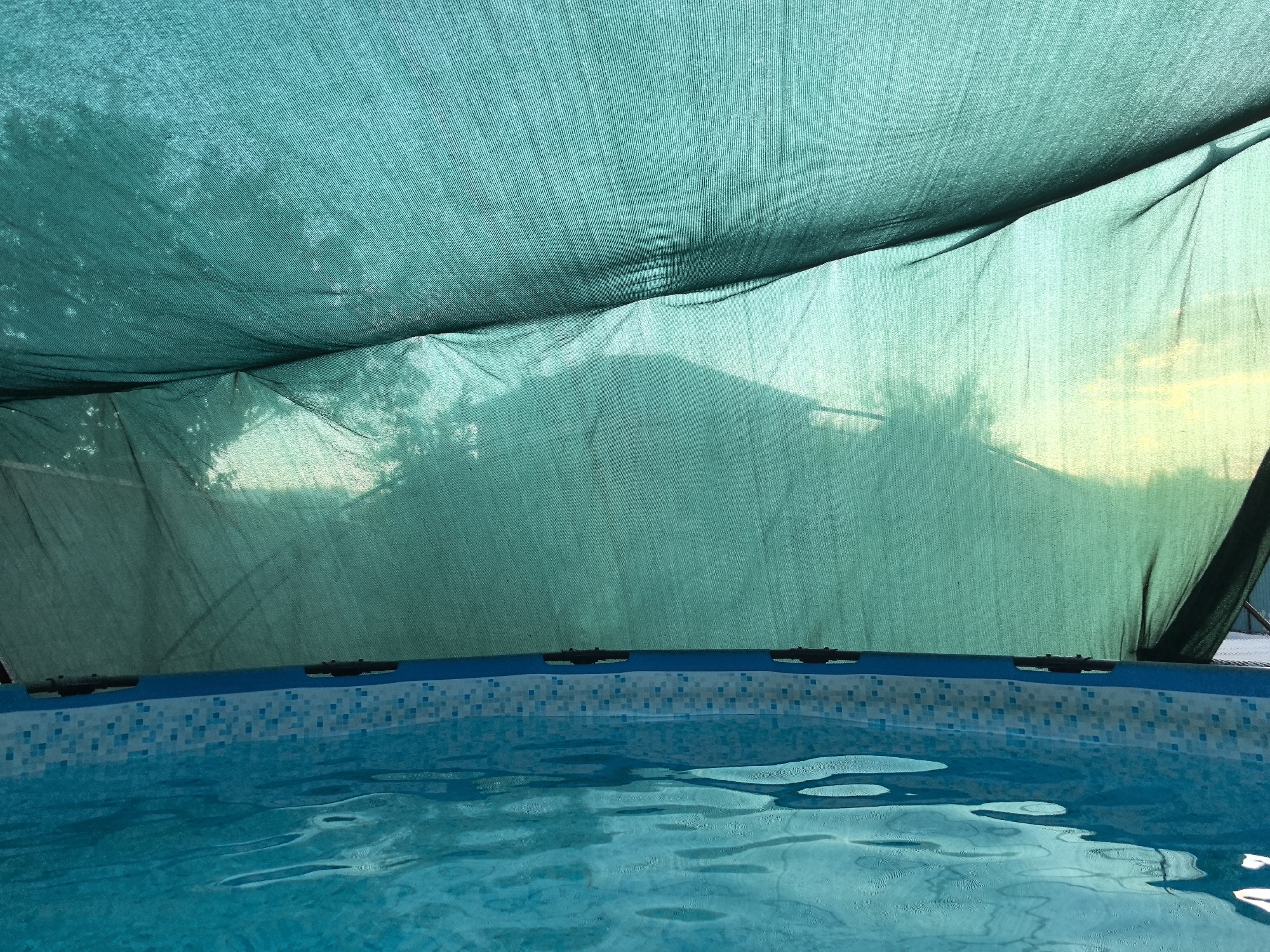 The image depicts an above-ground swimming pool filled with turquoise water, reflecting sunlight and casting dappled shadows. The pool has a patterned liner around its rim. Above the pool, a large green shade cloth is stretched, creating a canopy that filters the sunlight and casts a silhouette of trees and possibly a building in the distance. The sky is visible through the shade cloth, transitioning from a bright yellow near the horizon to a lighter tone. The scene appears to be outdoors, likely in a residential setting. The location is not specified in the image or its accompanying information.