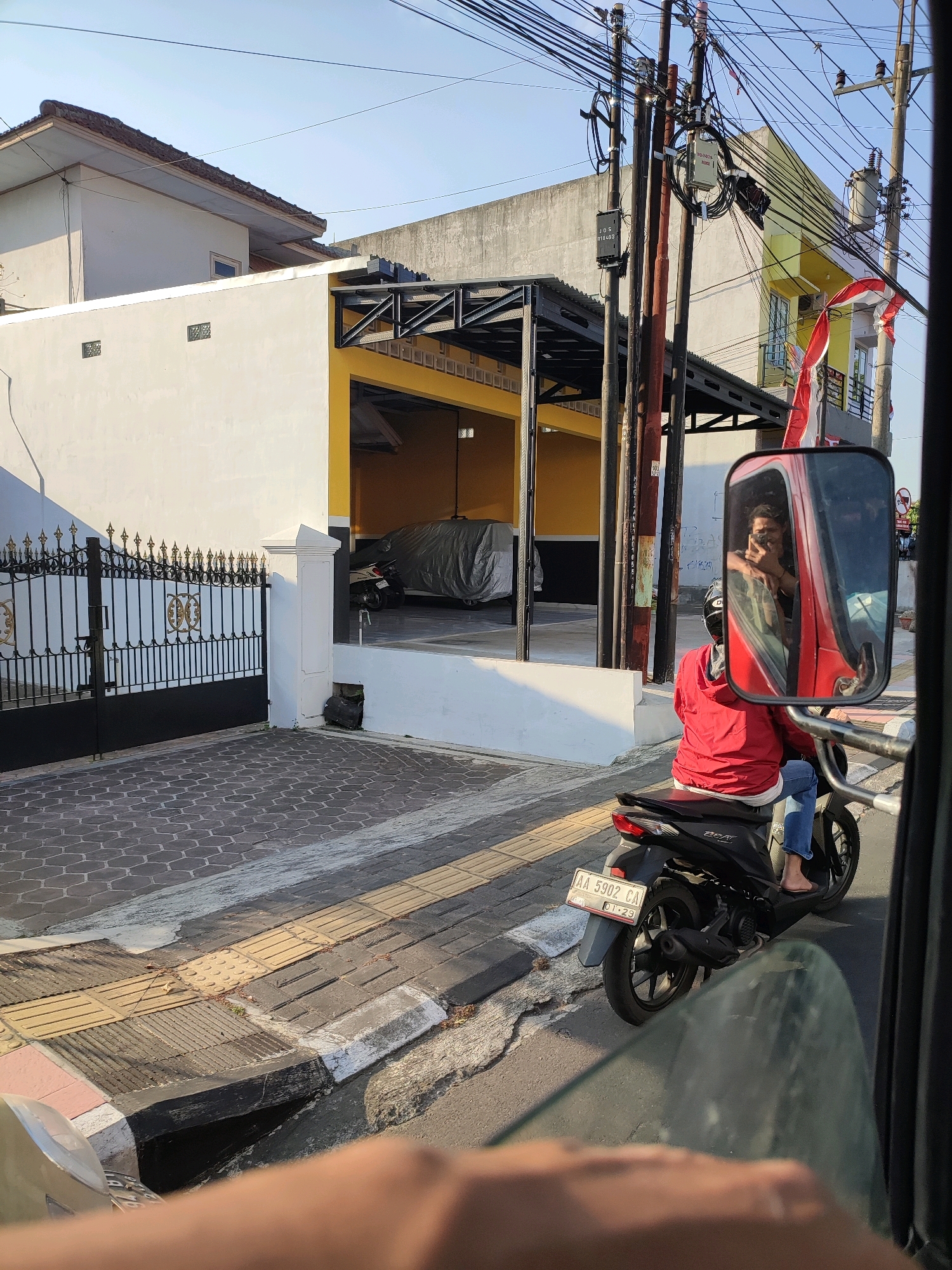 The image depicts a street scene in the city of Magelang, Indonesia, captured during daylight hours. In the foreground, the perspective suggests the shot is taken from inside a vehicle, with a blurry hand and a portion of a car window visible. To the right, a motorcycle is prominently featured, viewed from the rear. The rider, wearing a red jacket and blue jeans, is seated on the black motorcycle, which has a license plate reading "AA 5902 CA" and "01-29". The rider's face is reflected in the side mirror of a red vehicle, where they appear to be taking a photograph with a phone.
The background reveals a building with a white facade and a yellow section, possibly a garage or covered parking area, with a black metal awning. A black wrought-iron gate with ornate details stands to the left of the building, leading to a paved courtyard. A motorcycle covered in a gray tarp is partially visible inside the covered area. Overhead, a dense network of electrical wires and poles crisscrosses the sky, indicating an urban environment. A partial view of a building with yellow siding and a red and white Indonesian flag is also visible to the right, suggesting the presence of more structures and activity. The overall setting is characterized by a paved street, sidewalks, and typical urban infrastructure.