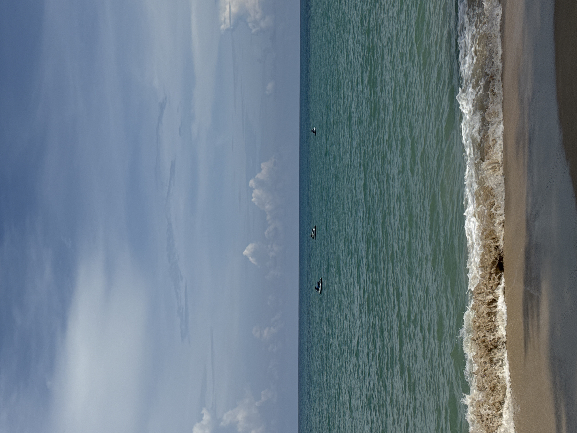 The image depicts a serene ocean scene at Bang Tao Beach in Ban Bang Thao, Thailand. The horizon is clearly defined, separating the tranquil turquoise water from a sky filled with scattered, fluffy white clouds against a backdrop of lighter blue. A gentle wave, tinged with sandy brown sediment, breaks onto the shoreline, which consists of light-colored sand. In the distance, three jet skis are visible on the water. One is positioned to the left of center, with a person on it, while two others are further to the right. The overall impression is one of peaceful coastal recreation.