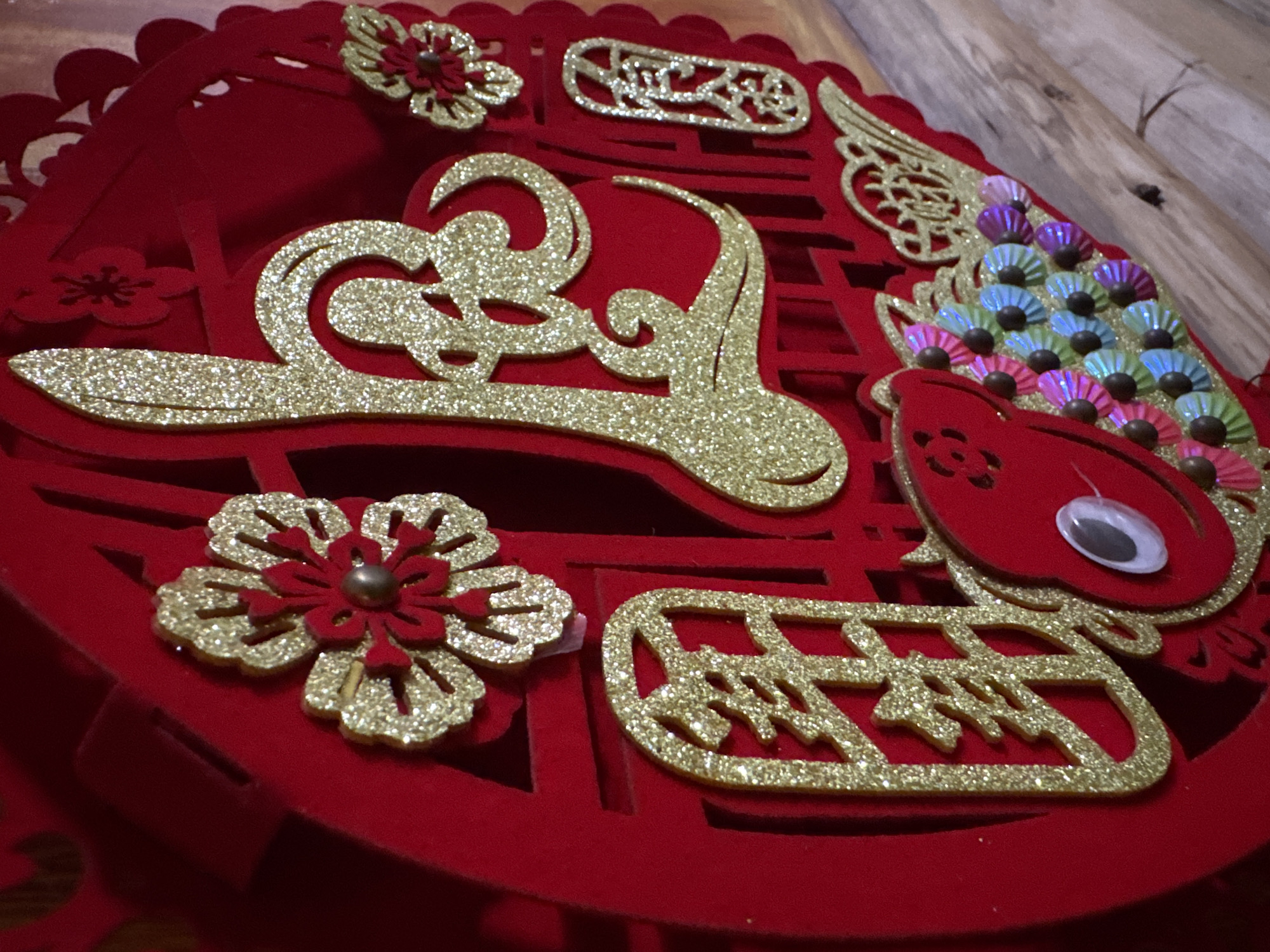 The image features a close-up view of a red, decorative craft object. The object appears to be made of red felt or similar fabric and has intricate cut-out designs. Adorned with glittery gold embellishments shaped like flowers, stylized lettering, and swirling patterns. Notably, there is a red bird-shaped element with what appears to be a googly eye and a fan-like tail made of iridescent, colorful scales. The overall design suggests a celebratory or festive theme. The background is out of focus, showing wooden planks. The location in Mèo Vạc Commune, Vietnam is not discernable from the image alone.