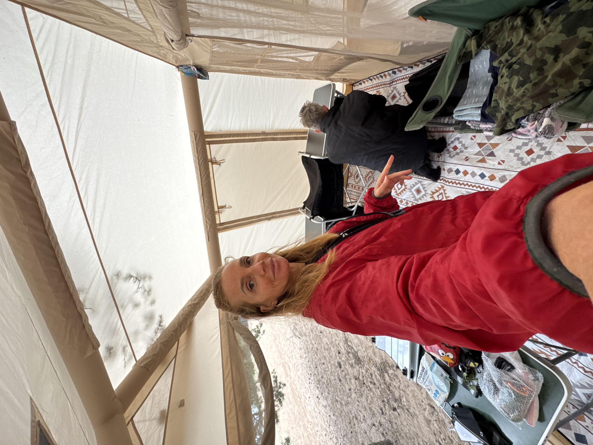 The image captures an indoor scene within a tan canvas tent, likely in the city of Ozerki (Krasnodolinskaya), Russia. A blonde woman wearing a red jacket is in the foreground, taking a selfie and smiling at the camera. Her left hand is extended, with her index finger pointing towards the camera. In the background, a person with grey hair, wearing a black jacket, is seen from the back, crouching near a foldable table. The tent's interior features a patterned rug on the ground, a foldable table with various items on it, including what appear to be hygiene products and a red Angry Birds plush toy. Folding chairs and folded clothing, including a camouflage garment, are also visible within the tent. The environment outside the tent, visible through an opening, appears to be sandy terrain with sparse vegetation.