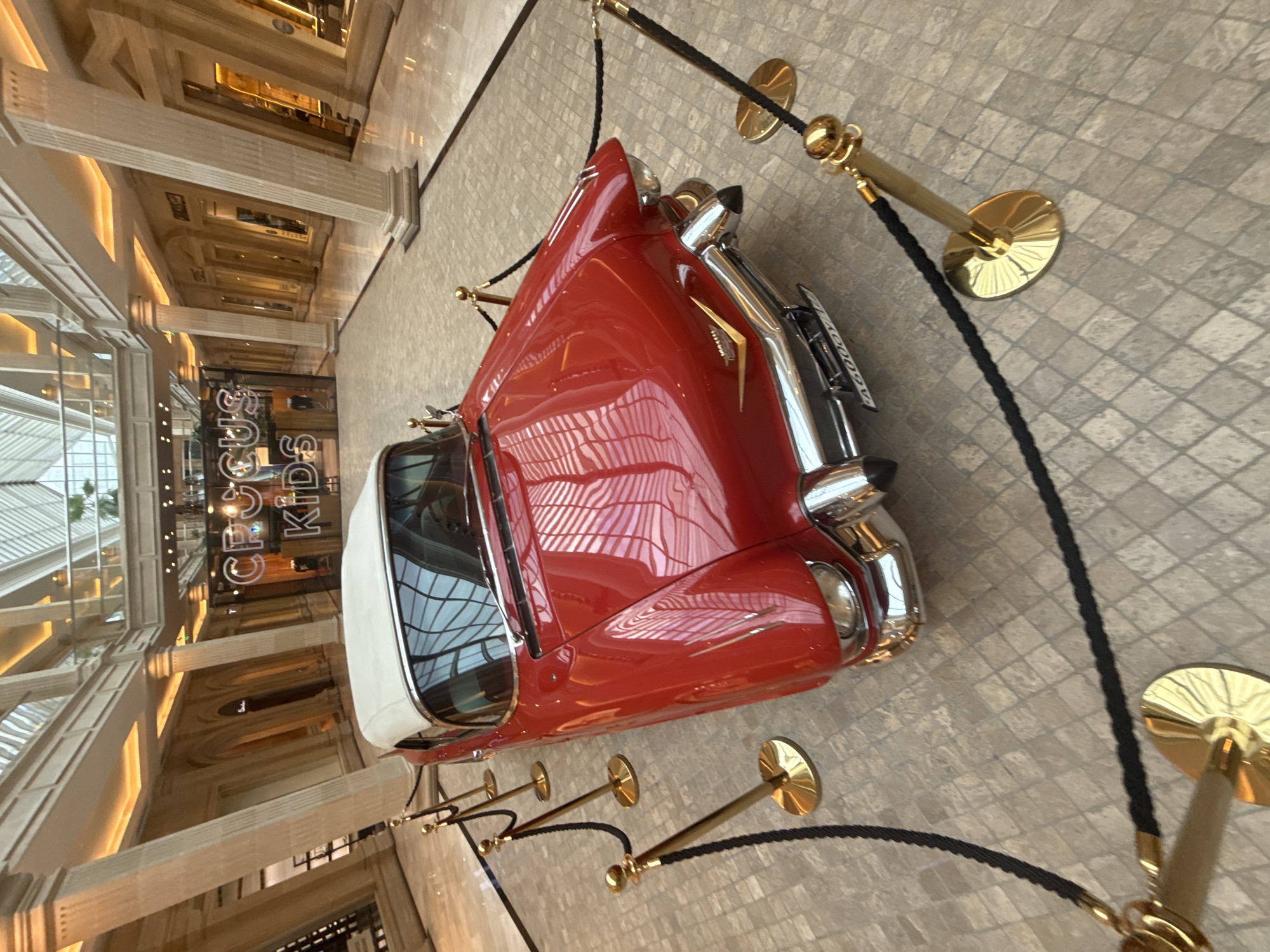 A vintage red convertible Cadillac is displayed on a polished stone floor within a large, open, modern indoor space. The car has a white soft top. Gold stanchions with black ropes are arranged around the car, indicating a cordoned-off display area. In the background, there are shops with signage, including "CROCUS KIDS." The architecture suggests a luxury shopping mall with tall columns and a high ceiling with natural light. The location is Crocus City Mall in the city of Khimki, Russia. There are no people visible in the image.