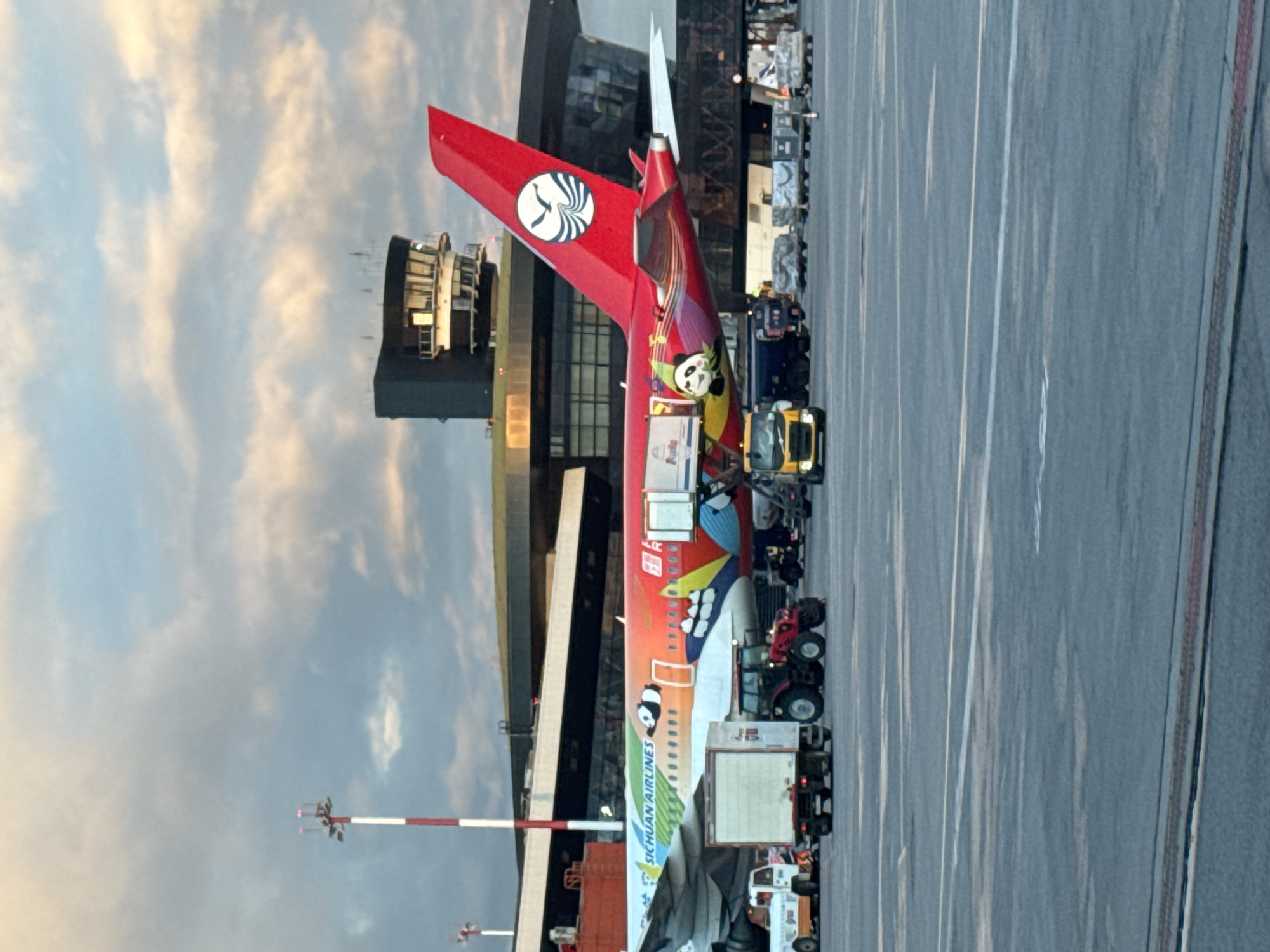 The image shows the tail section of a Sichuan Airlines aircraft at Sheremetyevo International Airport, outside Khimki, Russia. The aircraft is decorated with cartoon panda imagery. Ground service vehicles surround the plane. The sky is overcast. An airport building is visible in the background.