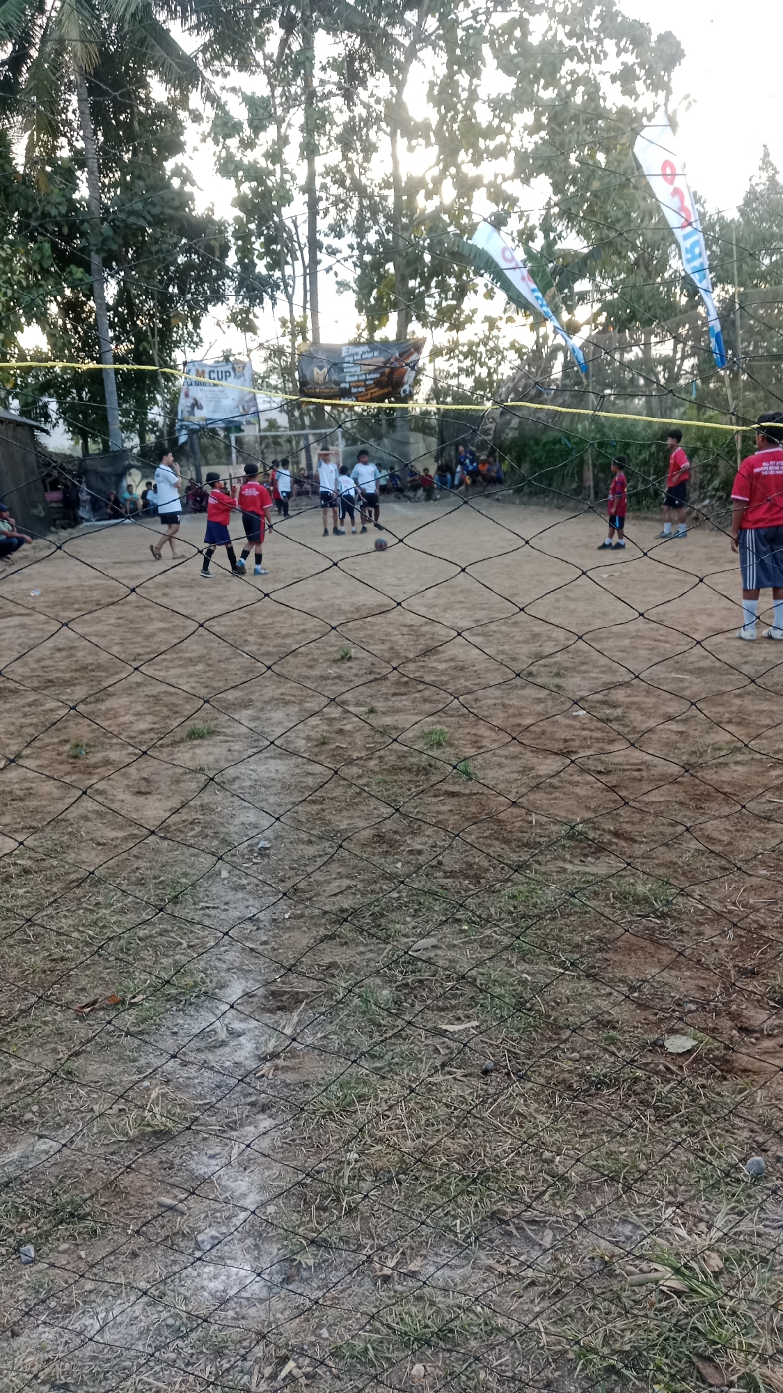 This image depicts an outdoor volleyball game taking place on a dirt court in Karangharjo, Indonesia. Several young people, likely teenagers, are actively participating in the game, dressed in athletic attire. Some are wearing red shirts and black shorts, while others are in white shirts and dark shorts. A volleyball is visible on the court, suggesting an ongoing match. In the background, trees and foliage frame the scene, and banners with text are strung up, indicating a local event or tournament. The presence of a volleyball net divides the court. Spectators can be seen in the background, seated or standing on the periphery. The overall atmosphere appears to be casual and community-oriented, with the game being played in a rustic, outdoor setting.