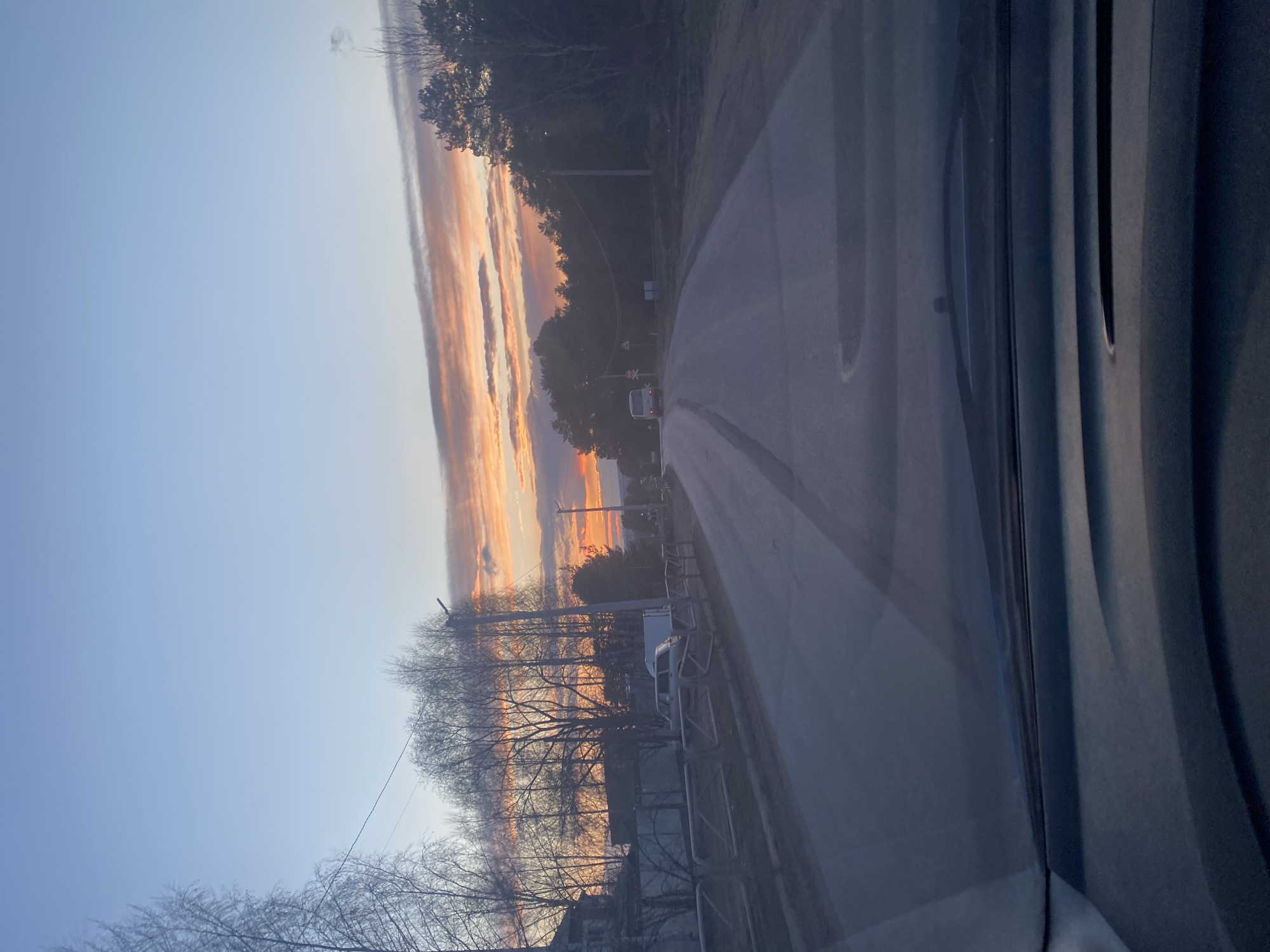The image captures a rural or semi-rural road scene at either sunrise or sunset, viewed from inside a vehicle in Chaikovsky, Russia. The sky dominates the left side of the frame, showcasing a clear blue transitioning into a spectacular display of orange, pink, and yellow hues along the horizon, reflecting off elongated cloud formations. This suggests a serene and colorful dawn or dusk.
The road, appearing paved and possibly wet in places, stretches into the distance, with a gentle curve to the right. On both sides of the road, various trees are visible; many deciduous trees have bare branches, indicating late autumn or early spring. Evergreen trees are also present, particularly on the left side. Utility poles with overhead lines run alongside the road.
Further down the road, a white van or small bus is visible, heading away from the viewer, with its taillights illuminated. Near this vehicle, faint road signs, possibly indicating a railroad crossing with a distinctive 'X' shape, can be observed. The right side of the image reveals part of the vehicle's interior, including the dark frame of a windshield or side window and what appears to be a dashboard element, indicating the photo was taken from within a car.
The overall impression is one of peaceful travel during a beautiful time of day, with the vibrant sky being the most prominent and aesthetically pleasing feature. There is no visible text in the image.