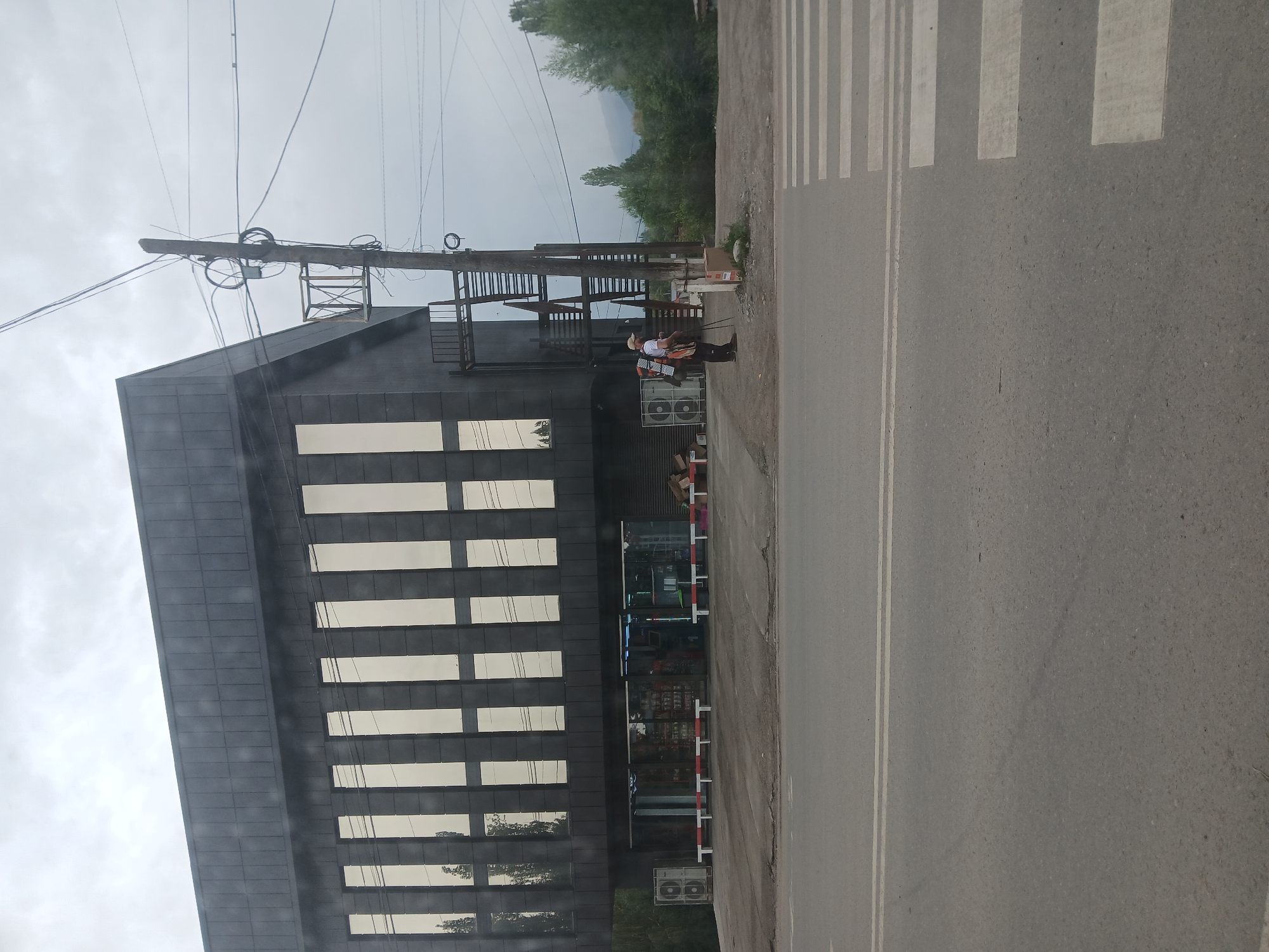 The image shows a modern, dark-colored building with a row of tall, reflective windows on its upper floor. The building appears to be a commercial establishment, with a glass storefront on the ground level displaying merchandise. In front of the building, there is a paved area with a crosswalk visible on the right side of the frame. A utility pole with numerous wires stands to the right of the building. Two individuals are present near the utility pole. One appears to be a person with a backpack and a hat, possibly using a tripod or camera equipment. Another person is standing nearby. The environment suggests a cloudy day in a location with some greenery and distant mountains, consistent with Ak-Suu, Kyrgyzstan.