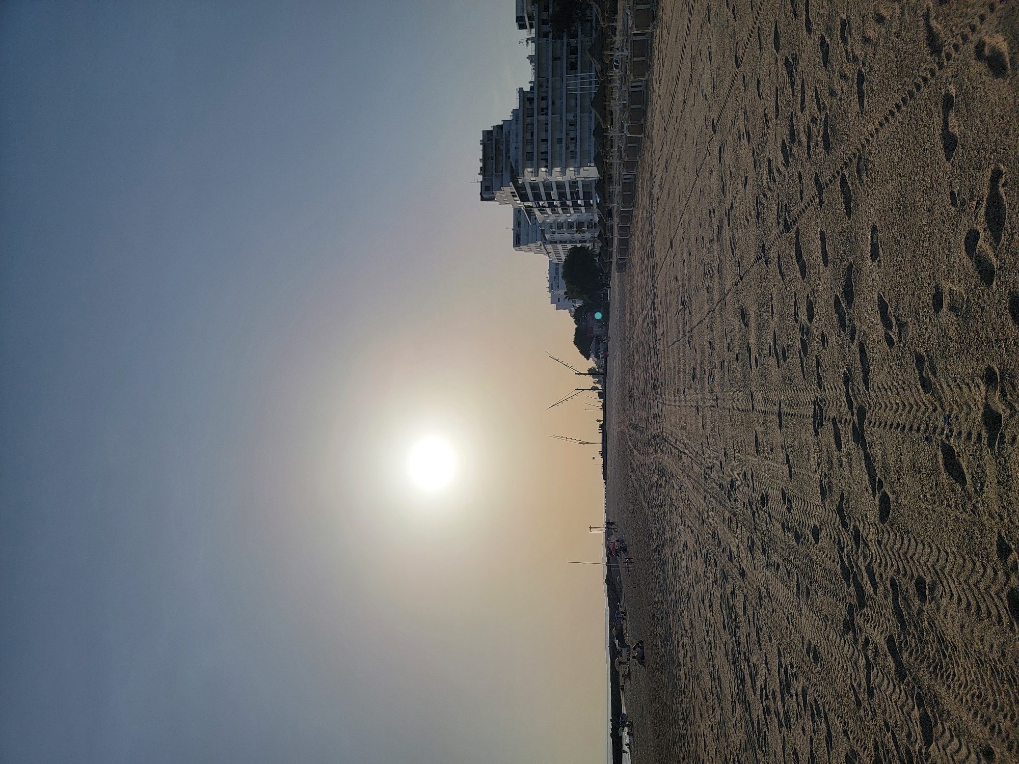The image captures a wide sandy beach in Quarteira, Portugal, under a bright, hazy sun. The foreground is dominated by the texture of the sand, marked with numerous footprints and tire tracks.
In the middle ground, a line of empty lounge chairs and thatched umbrellas stretches along the edge of the beach. Beyond them, several people are scattered across the sand, some sitting and others walking. In the distance, a low-lying barrier of dark rocks or a breakwater extends into the sea.
Further back, a row of modern, white apartment buildings lines the coast, interspersed with trees. The buildings appear to be multi-story and architecturally varied. Tall, slender lampposts are visible along the promenade in front of the buildings.
The sky is a pale blue, transitioning to a lighter, almost white haze near the horizon due to the sun's brightness. The overall impression is a peaceful, late afternoon or early morning scene on a popular beach resort.