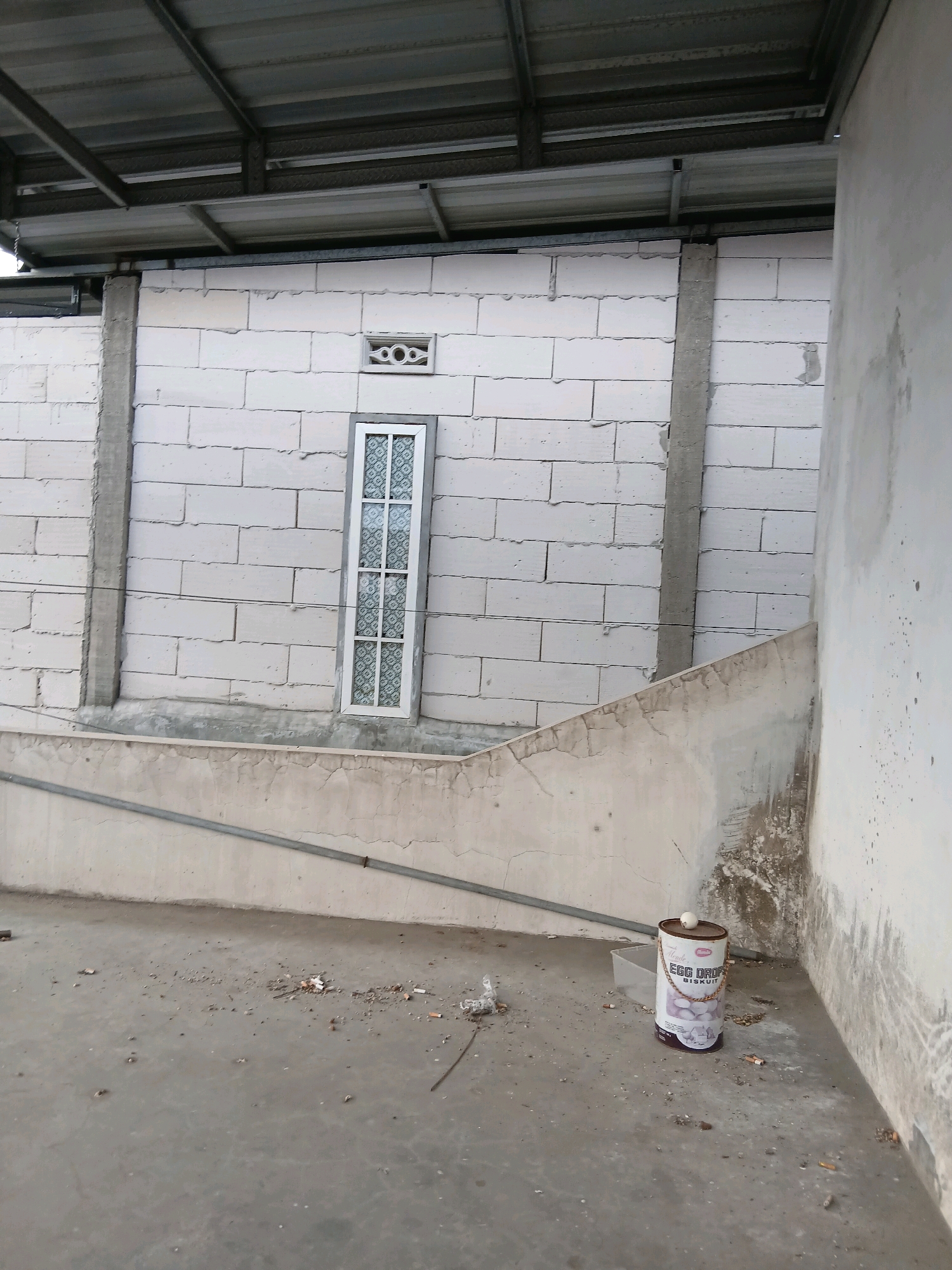 The image shows a construction site or unfinished building in Tenggarang, Indonesia. The main features include a white brick wall with a narrow, tall window, a corrugated metal roof supported by metal beams, and a concrete floor littered with debris. A metal pipe runs horizontally across the wall. In the foreground, a cylindrical tin labeled "EGG DROP BISCUIT" stands on the floor. The overall impression is one of a functional, outdoor space undergoing development or renovation.
