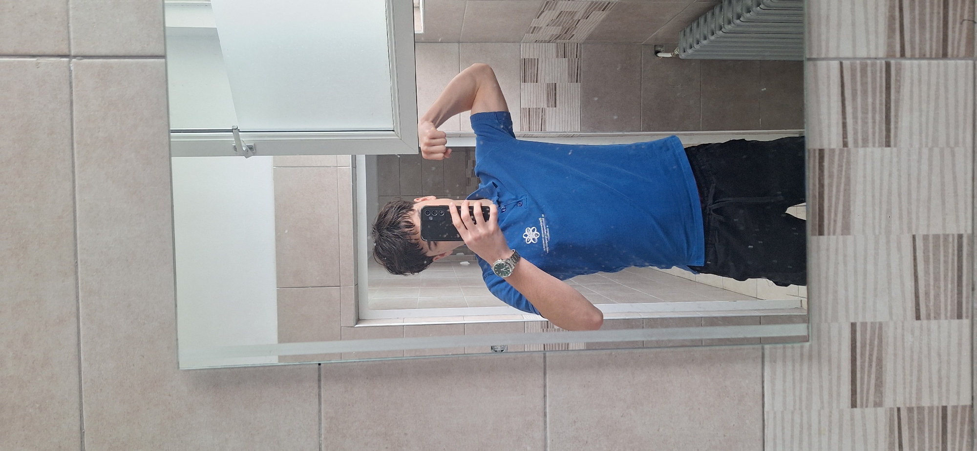 The image depicts a person taking a selfie in a bathroom mirror. The person is wearing a blue polo shirt with a logo on the left chest and dark-colored shorts. A watch is visible on their left wrist. They are holding a smartphone in front of their face. The bathroom has beige tiled walls and a window on the left. In the background, a radiator and patterned tiles can be seen. Based on prior information, it is presumed that the location is Burgas, Bulgaria.