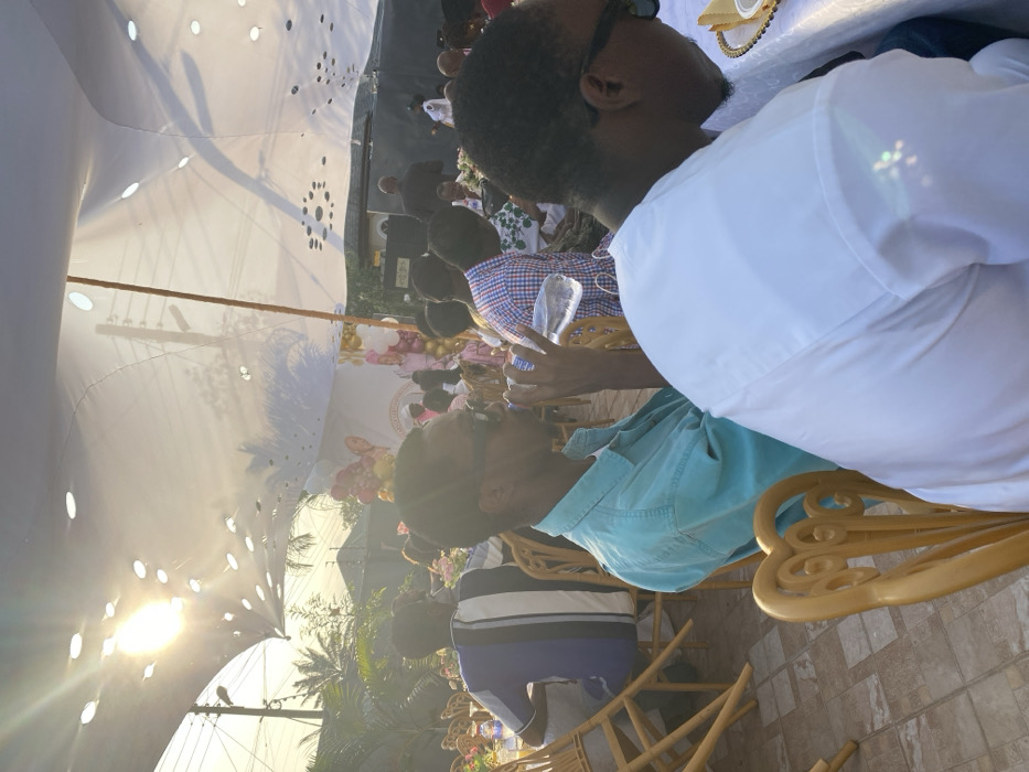 An outdoor event unfolds beneath a white tent with circular cutouts allowing sunlight to filter through. Attendees are seated on gold chairs, dressed in various attire including a blue button-down shirt, white shirt, and blue striped shirt. A man in sunglasses and a white shirt is prominently visible. Behind the crowd, there is greenery and a decorated backdrop. Balloons and flowers add a festive touch to the event.