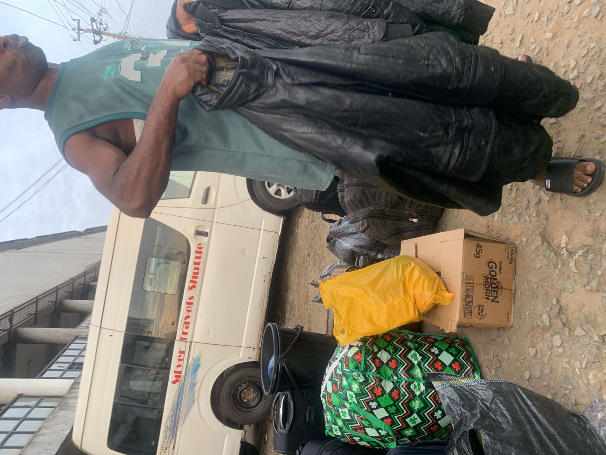 A medium shot captures a man in Elelenwo, Nigeria, standing in front of a white shuttle van branded "Silver Travels Shuttle." He is holding several dark-colored jackets, one of which appears to be made of leather. The man wears a sleeveless green shirt and black sandals. The foreground of the image is filled with various bags, including a green and white patterned bag, a bright yellow plastic bag, a black bucket, and several black backpacks and bags. A cardboard box with "GOLDEN MORN" branding is also visible among the luggage. The ground is rough and appears to be dirt or gravel. The background shows a building with a covered walkway and what looks like overhead wires and a utility pole. The sky is overcast.