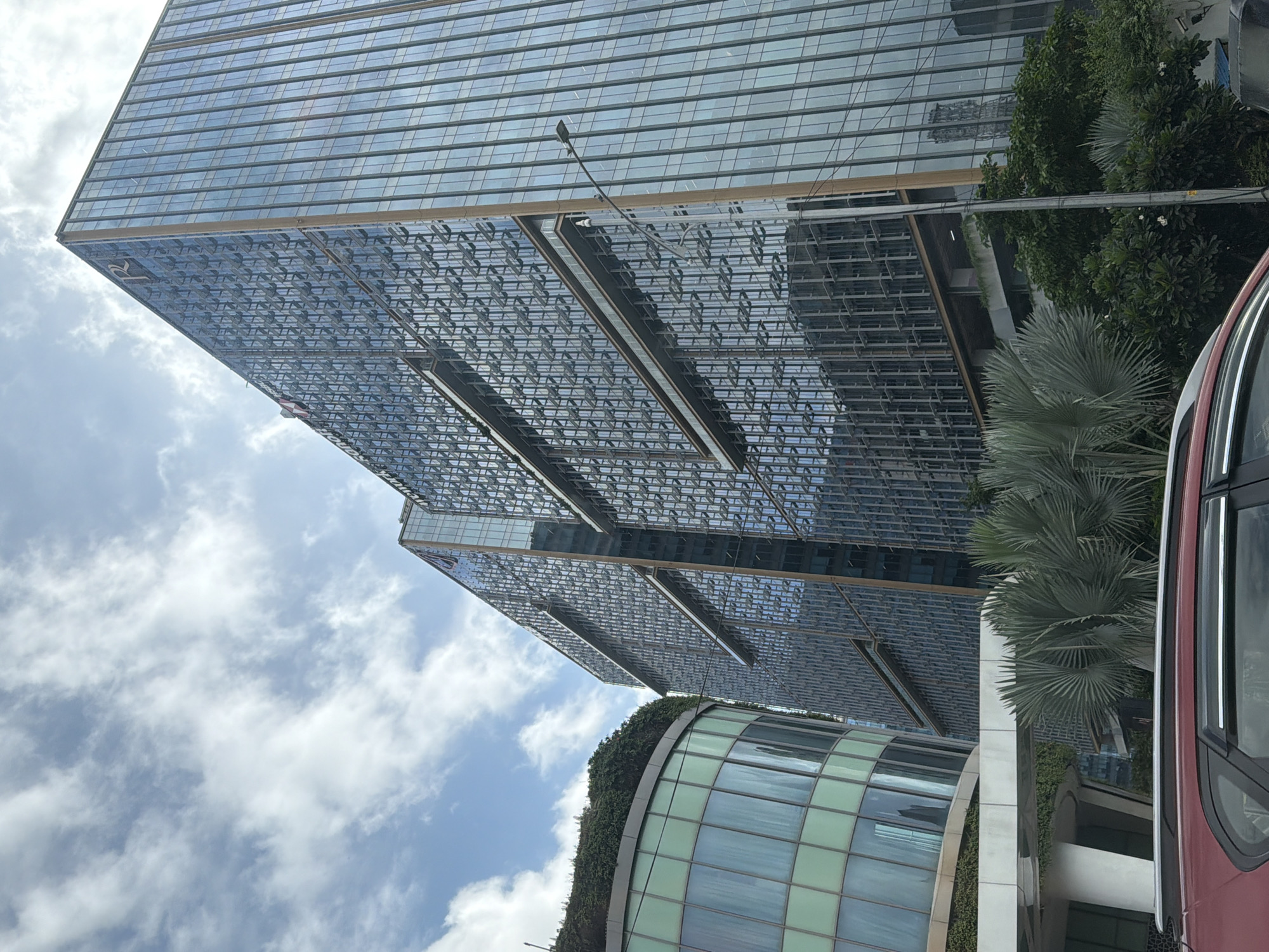 The image captures a modern cityscape featuring a large, glass-fronted building in Hyderabad, India, likely an office or commercial complex. The building is characterized by its extensive use of reflective glass panels and a warm-toned metal framework. A distinctive "R" logo is visible at the top of the building, possibly indicating the brand or company housed within. Adjacent to this structure, a curved building with green tinted glass panels and a lush green roof adds architectural contrast. In the foreground, the roof and partial side of a red car are visible, suggesting a street-level perspective. The scene is set against a backdrop of a cloudy sky, with palm trees and other greenery adding a touch of nature to the urban environment. The overall impression is one of contemporary architecture and urban development in Hyderabad.