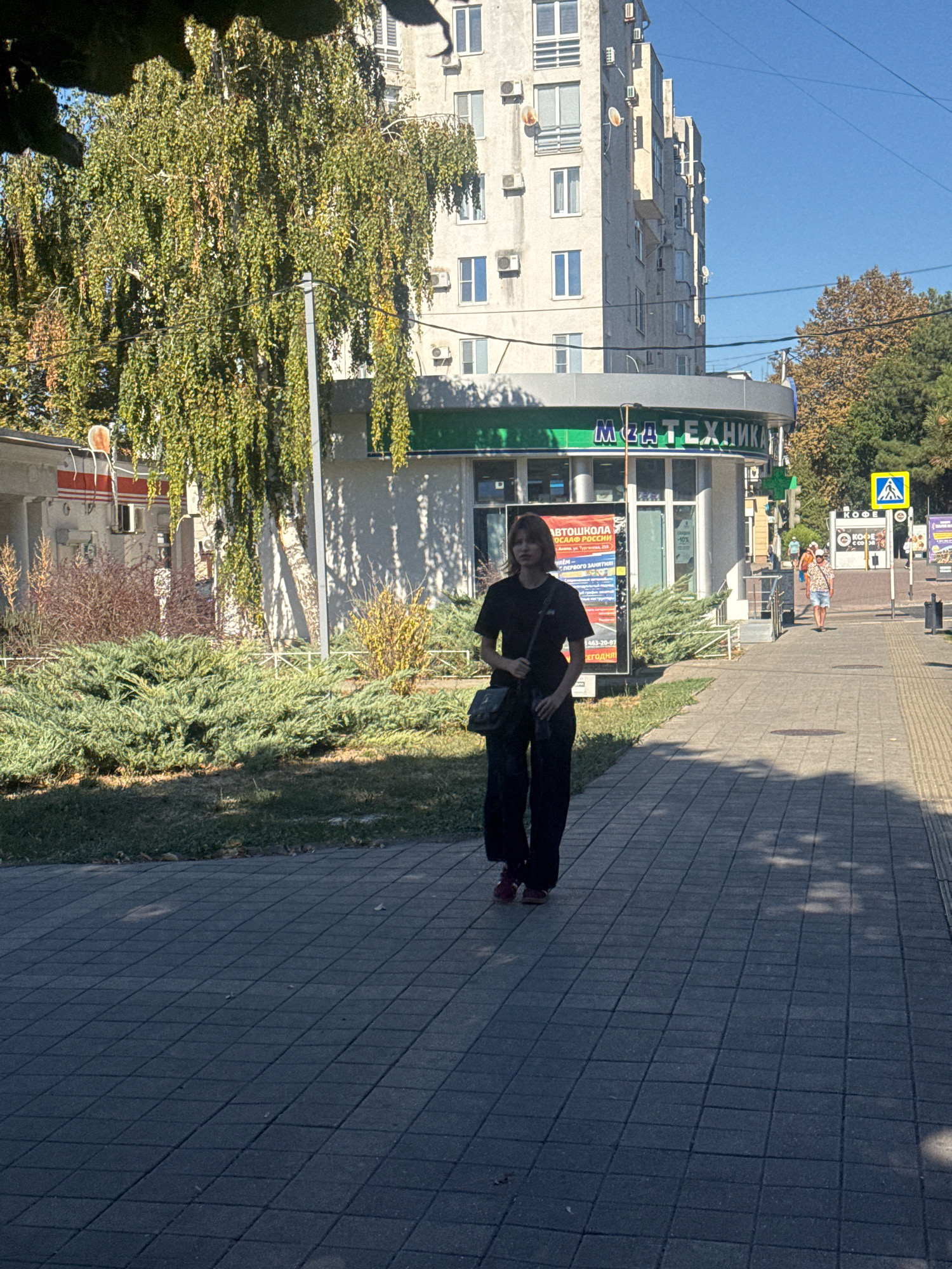 The image shows a young person wearing a black t-shirt and dark pants walking on a paved sidewalk. They have a dark-colored bag slung across their body. The person is walking towards the viewer. Behind them, there is a building with several floors and air conditioning units visible on its facade. A green awning with Russian text is above the entrance of a business on the ground floor. To the left of the building, there are bushes and a tall tree with drooping branches. Further down the sidewalk, other pedestrians are visible, as well as a yellow pedestrian crossing sign. The sky is clear and blue, and the sun is casting shadows on the sidewalk. The overall scene depicts a typical urban environment. The business on the ground floor of the building has the name "МЯТА ТЕХНИКА" written on its awning, which translates to "Mint Technique". There is also a sign for an "AUTOMOTIVE SCHOOL" and advertisements visible in front of the establishment. The presence of the name "Anapa" on one of the advertisements confirms the location as Anapa, Russia.