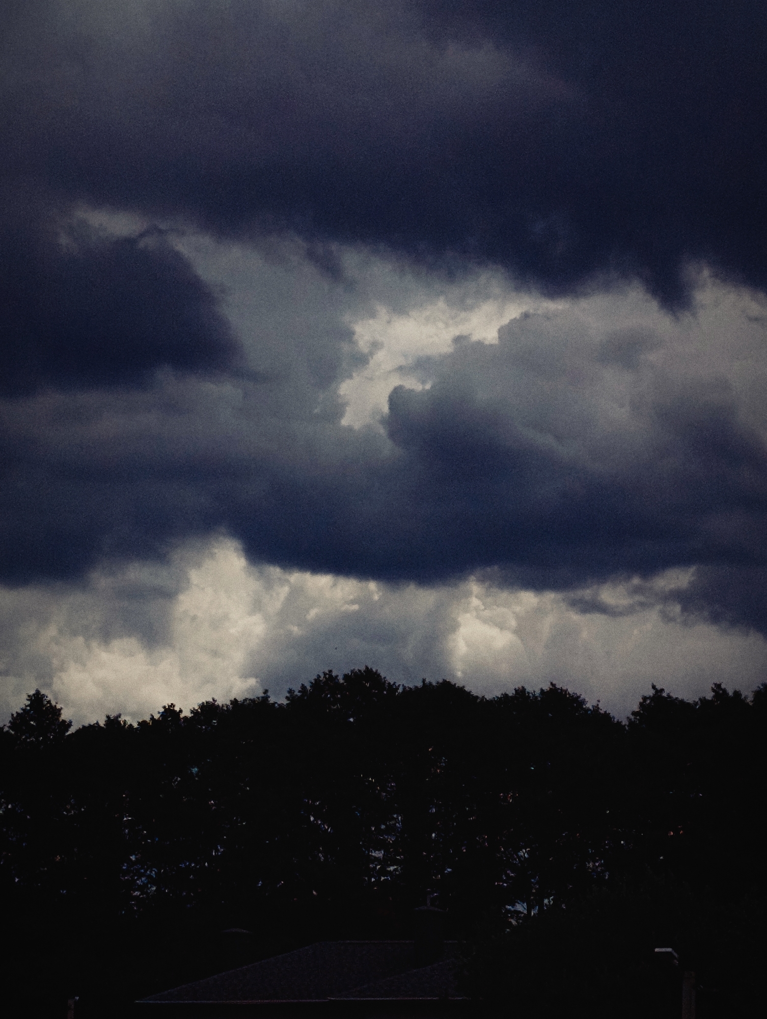 A dark, overcast sky looms above a silhouette of trees and a roofline in Minsk, Belarus. The clouds are heavy and turbulent, with some areas appearing brighter where light breaks through, suggesting a storm is approaching or has recently passed. The trees form a dense, dark band across the middle ground, obscuring whatever lies beneath them. The roofline at the bottom of the frame is also silhouetted against the sky. The overall impression is one of foreboding weather and muted light.