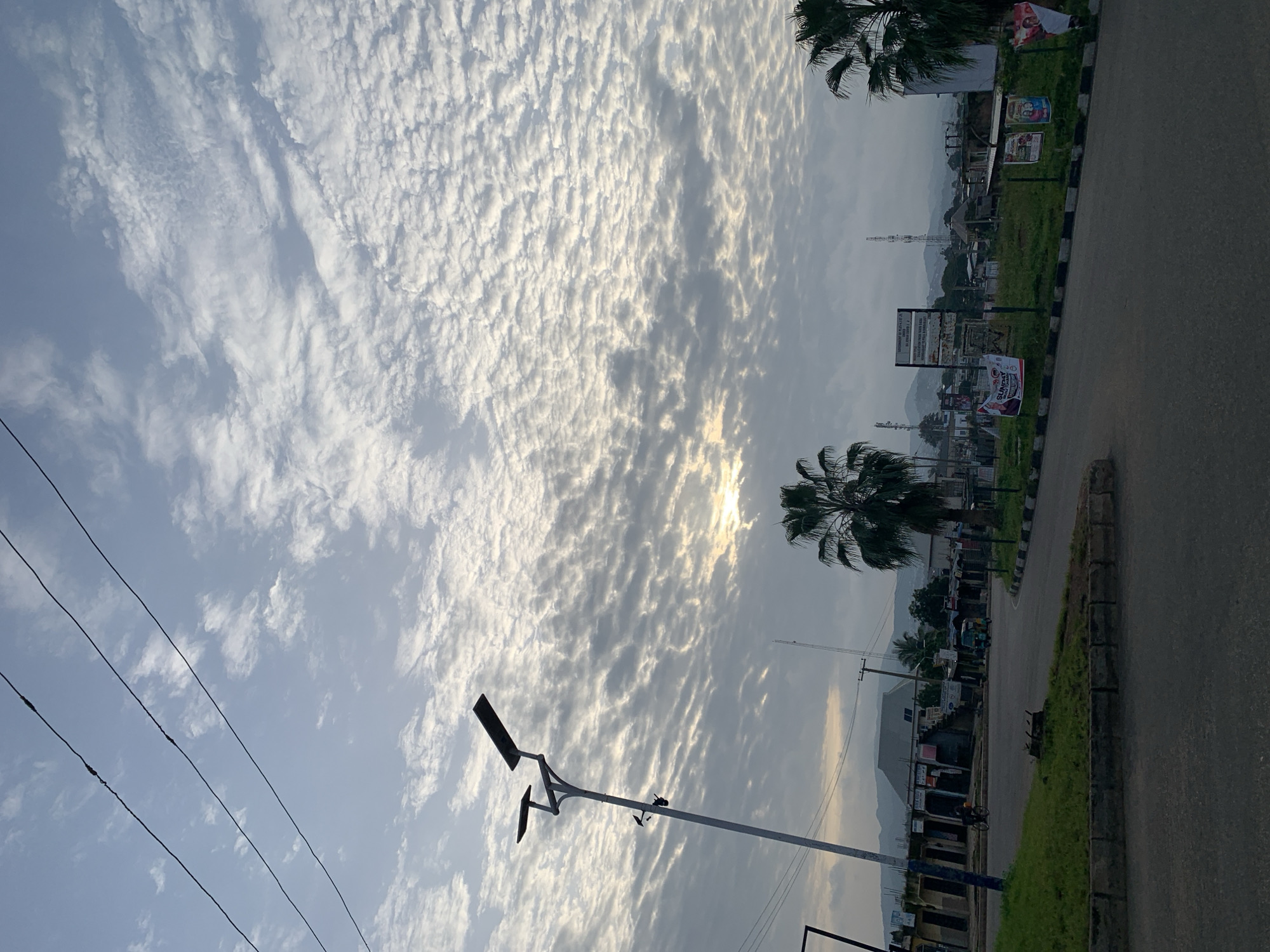 The image depicts an outdoor scene in Rafin Par, Nigeria, likely during early morning or late afternoon given the soft light. In the foreground, a paved road curves to the right, bordered by a grassy median strip with a stone curb. A modern street lamp with two solar panels stands on the left side of the road, its pole casting a shadow. In the background, several buildings with awnings and signs are visible, suggesting a commercial area. A man on a bicycle is moving away from the camera on the left, and a three-wheeled vehicle is stationary on the right side of the road. Palm trees are present, with one prominent tree in the middle ground and another on the far right. Communication towers are visible in the distance, along with a range of hills. The sky is filled with scattered, textured clouds, with the sun partially visible through them, creating a diffused light. Several posters are attached to poles in the grassy median, one of which features a portrait of a person. The overall impression is that of a quiet urban or semi-urban setting.