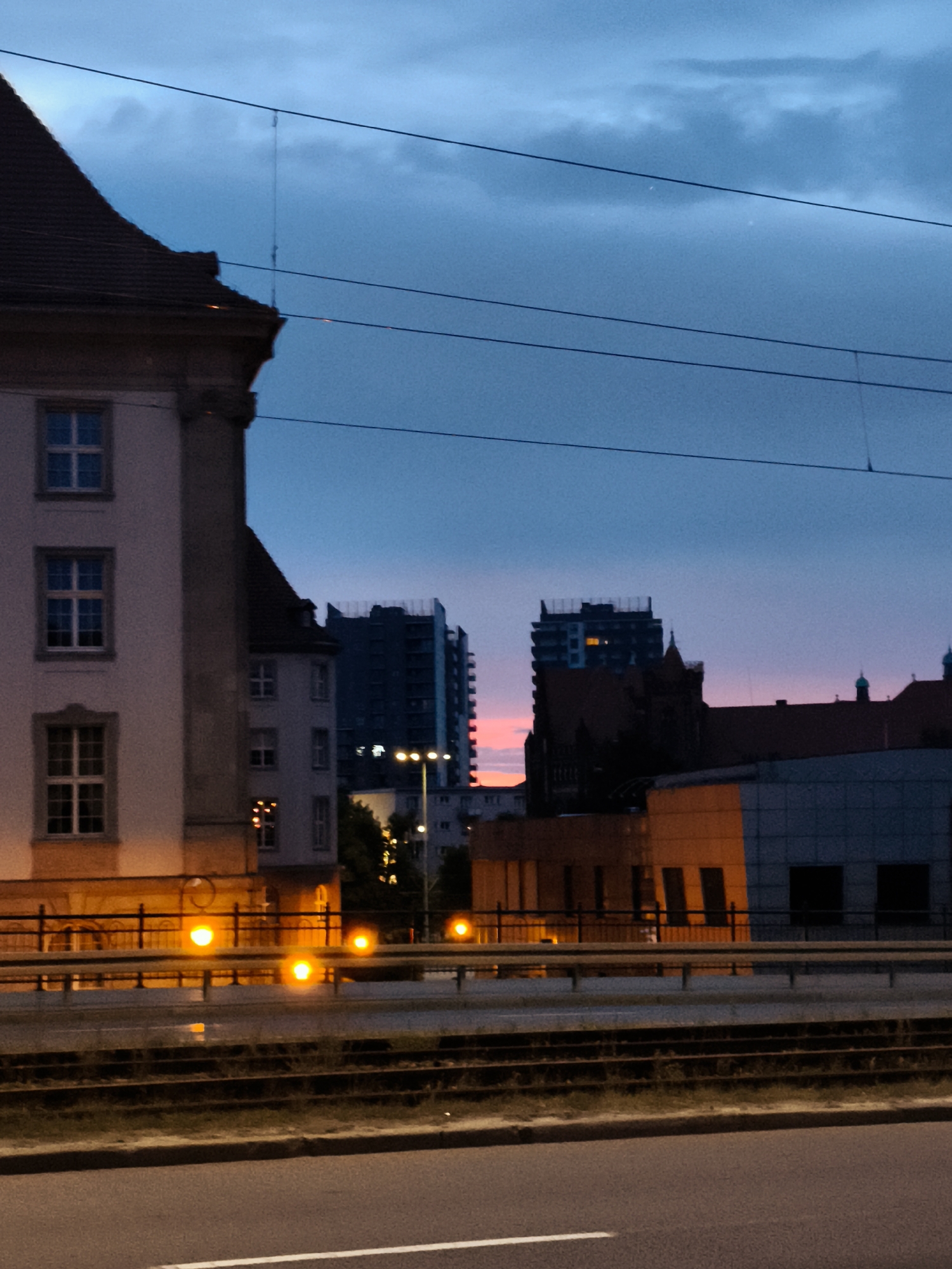 This is a vertical, eye-level, medium shot captured at dusk or dawn in the city of Gdańsk, Poland. The scene depicts a city street with a road in the foreground and a railway track behind it. In the middle ground, there is a pedestrian overpass with a black metal railing and several bright, yellow-orange lights illuminating the scene from street lamps. Behind the overpass, a multi-story building with multiple windows is visible, and further in the background, several tall buildings, including what appears to be a construction site or modern high-rise apartment complex, are silhouetted against the twilight sky. The sky itself is a deep blue, transitioning to a lighter hue near the horizon, with some scattered clouds. The overall atmosphere is urban and somewhat moody, typical of a city setting at the end or beginning of the day. No people are visible in the image.
