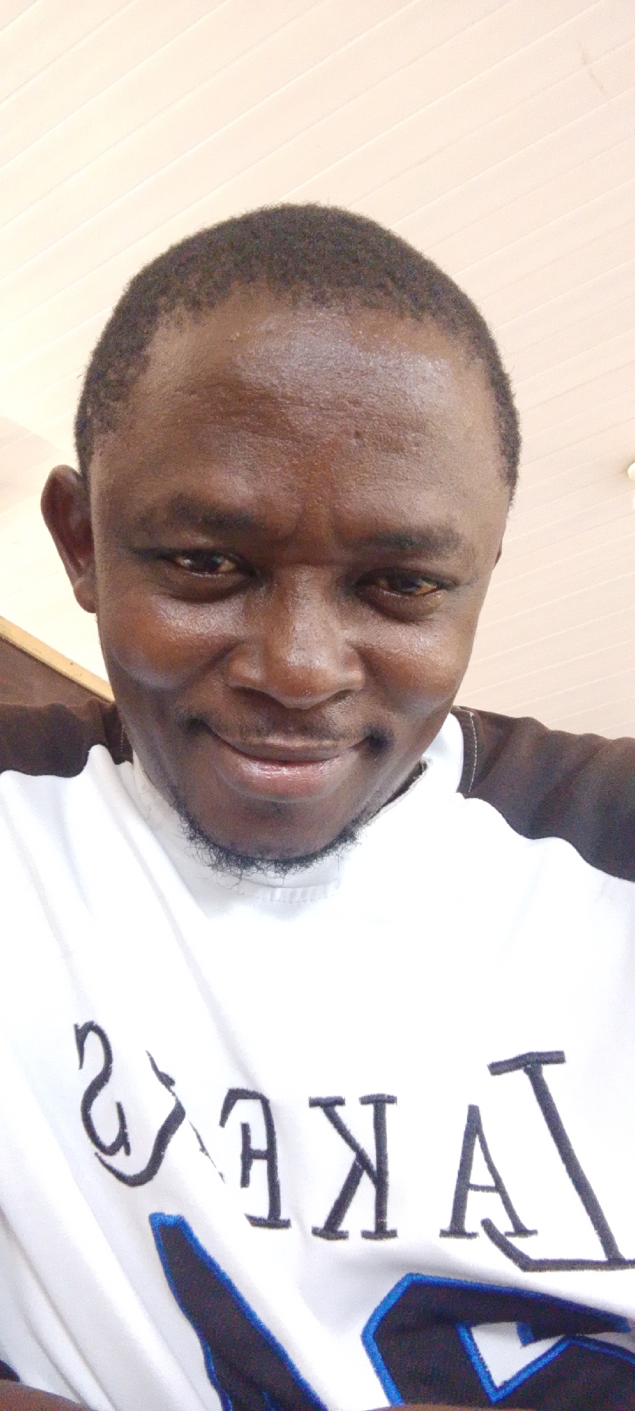 The image presents a close-up, selfie-style view of a dark-skinned man. He has short, dark hair and a light beard. He is wearing a white T-shirt with the word "LAKERS" printed on it. He appears to be indoors, with a light-colored ceiling visible in the background.
There is no indication of the city of Lokoja, Nigeria, in the image. No environmental context or features are present that would identify the location.
