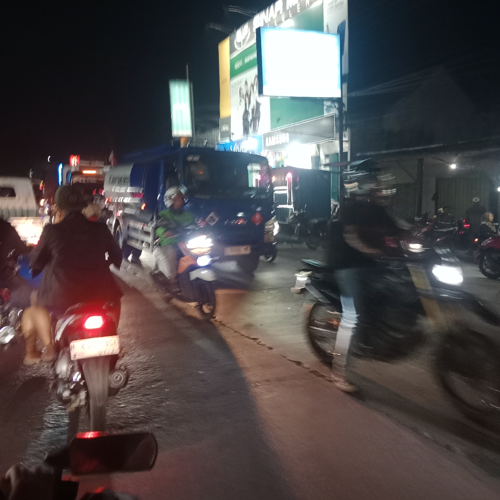 The image captures a night scene in Karangawen, Indonesia, characterized by traffic congestion. Several motorcycles and a truck are visible on a wet asphalt road. The lighting suggests a busy street with neon signs and illuminated advertisements in the background, including one for Samsung. One motorcycle in the foreground has a license plate that appears to be from Indonesia. The overall impression is one of a typical evening in a populated Indonesian town or city, with vehicles and pedestrians navigating through the nighttime conditions.