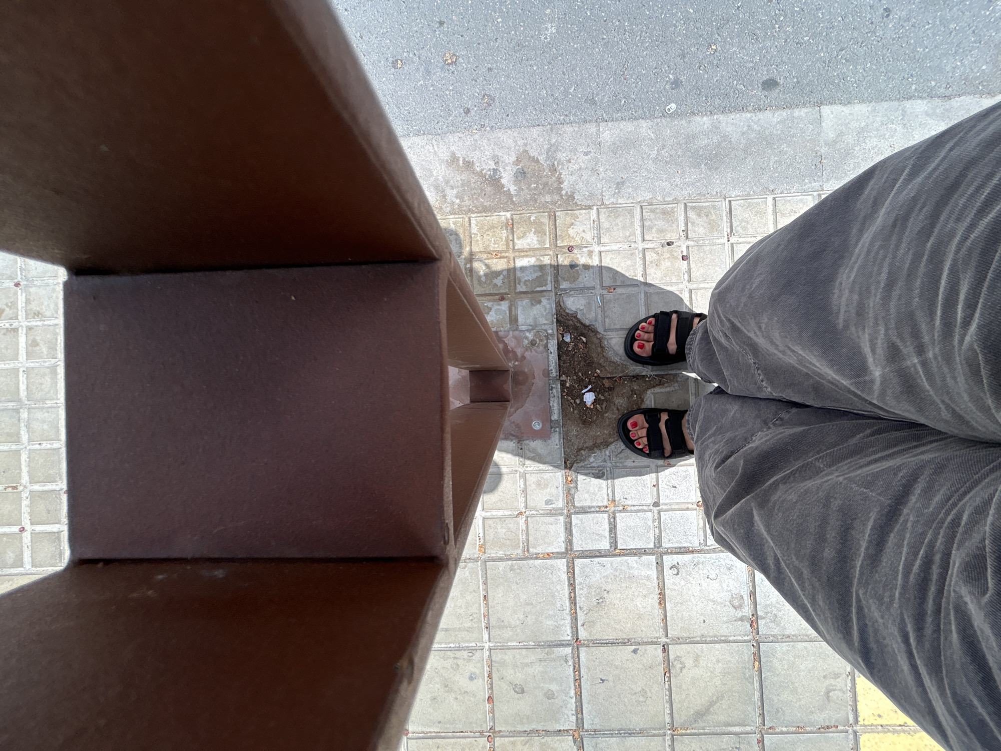 A first-person perspective shot shows a person's legs and feet standing on a sidewalk in Castelló De La Plana, Spain. The person is wearing gray corduroy pants and black sandals with red painted toenails. They are standing on a tiled sidewalk next to a brown, metallic structure. The tiles are light gray and appear weathered. To the right of the tiles is asphalt pavement. Sunlight casts a shadow of the metallic structure onto the sidewalk. There is some debris on the ground, including a small piece of white paper and some dried leaves.
