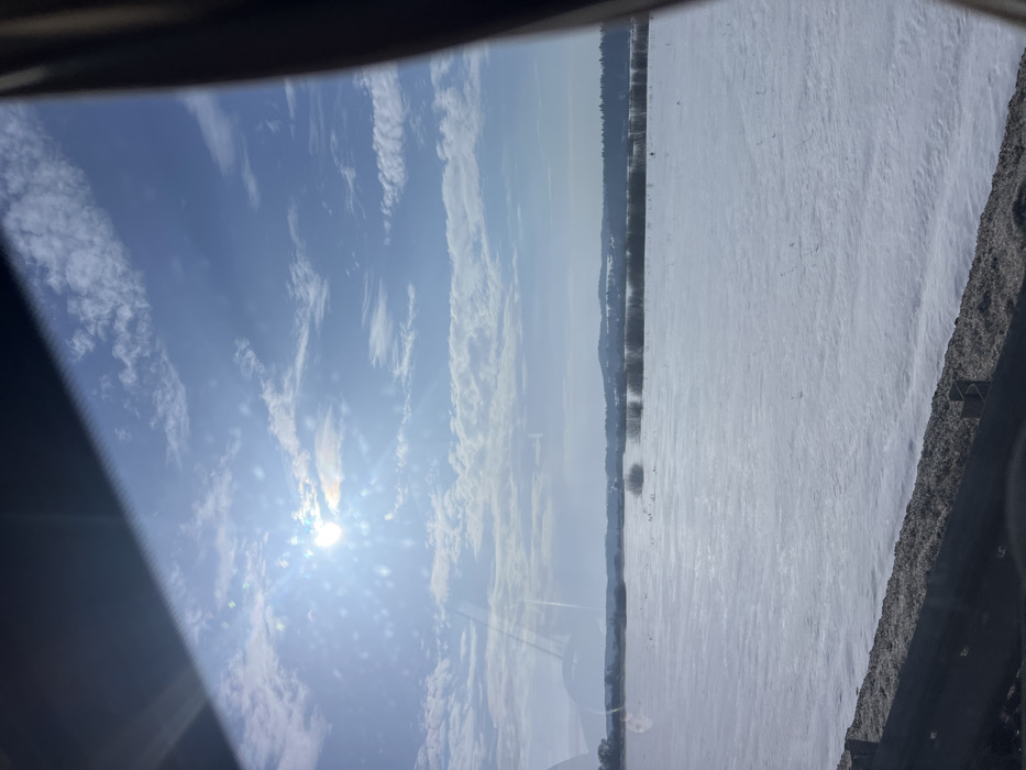 The image displays a winter landscape under a bright, clear blue sky with scattered white cumulus clouds, captured from inside a vehicle. The sun, a prominent luminous circle, is positioned in the upper-left quadrant, generating significant lens flare and glare across the upper half of the view. The foreground, partially obscured by the vehicle's interior elements, shows a dark, textured roadside embankment or barrier. Extending from the midground to the horizon is a vast, flat field covered uniformly in white snow, with sparse dark patches of vegetation visible. A distinct treeline of leafless deciduous trees forms a band across the middle distance, beyond which gently rolling, snow-covered hills are discernible. No specific individuals, animals, or structures are visible within the natural landscape. The scene is located in Chernovskoe, Russia.