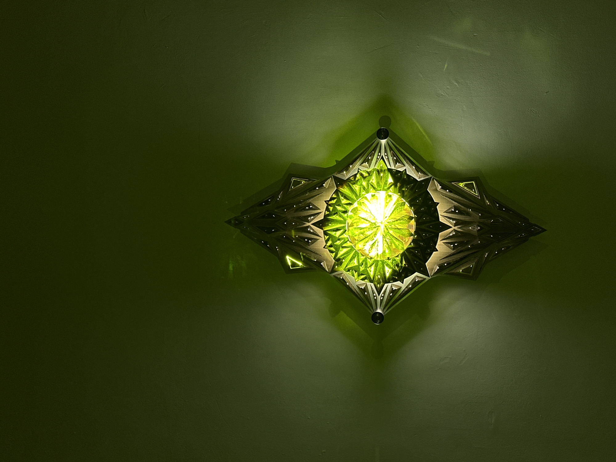 A low-angle shot captures a decorative, starburst-shaped wall sconce affixed to a green textured wall. The sconce, which is centered in the right portion of the frame, emits a bright, warm green light from its central circular area, casting intricate patterns of light and shadow onto the surrounding wall. The wall, predominantly green, fills the majority of the frame with variations in texture and tone due to the light source. No people or other objects are visible in the image. The location is not discernible from the visual information provided.