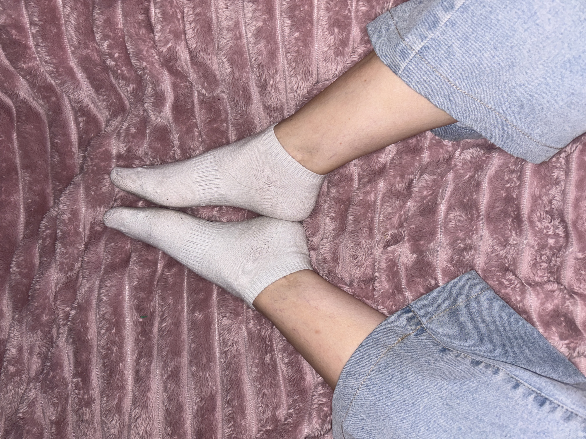 The image shows a close-up view of a person's legs and feet resting on a pink, plush blanket with horizontal ridges. The person is wearing white ankle socks and light blue jeans that are cuffed at the bottom. The lighting is dim, creating a soft glow on the fabric and skin. The scene appears to be indoors, possibly in a bedroom, with no visible indications of the specific city or country. The focus is on the texture of the blanket and the casual attire of the individual.