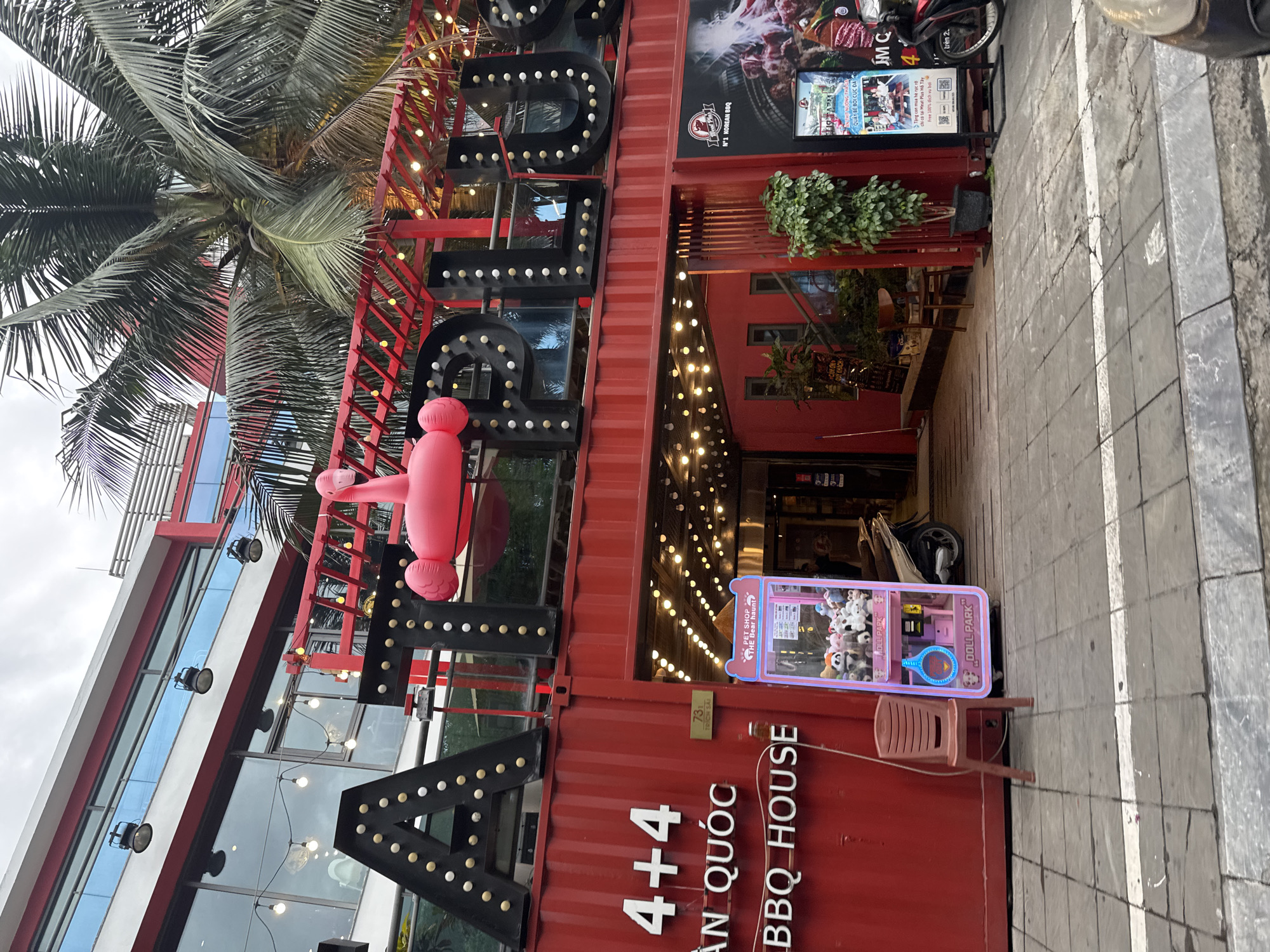 The image shows the exterior of a commercial establishment in Hanoi, Vietnam, identified by the text "BBQ HOUSE" and Vietnamese signage. The building is constructed from red shipping containers, with large black letters illuminated by light bulbs spelling out "AT PLUS" above the entrance. A pink inflatable flamingo is positioned above the letters. The entrance area is decorated with numerous string lights. To the left of the entrance, there is a red plastic chair and a pink "DOLL PARK" claw machine filled with stuffed animals. A motorcycle is parked near the right side of the entrance. A tall potted plant stands near the entrance. The ground is paved with square tiles. The sky is overcast.