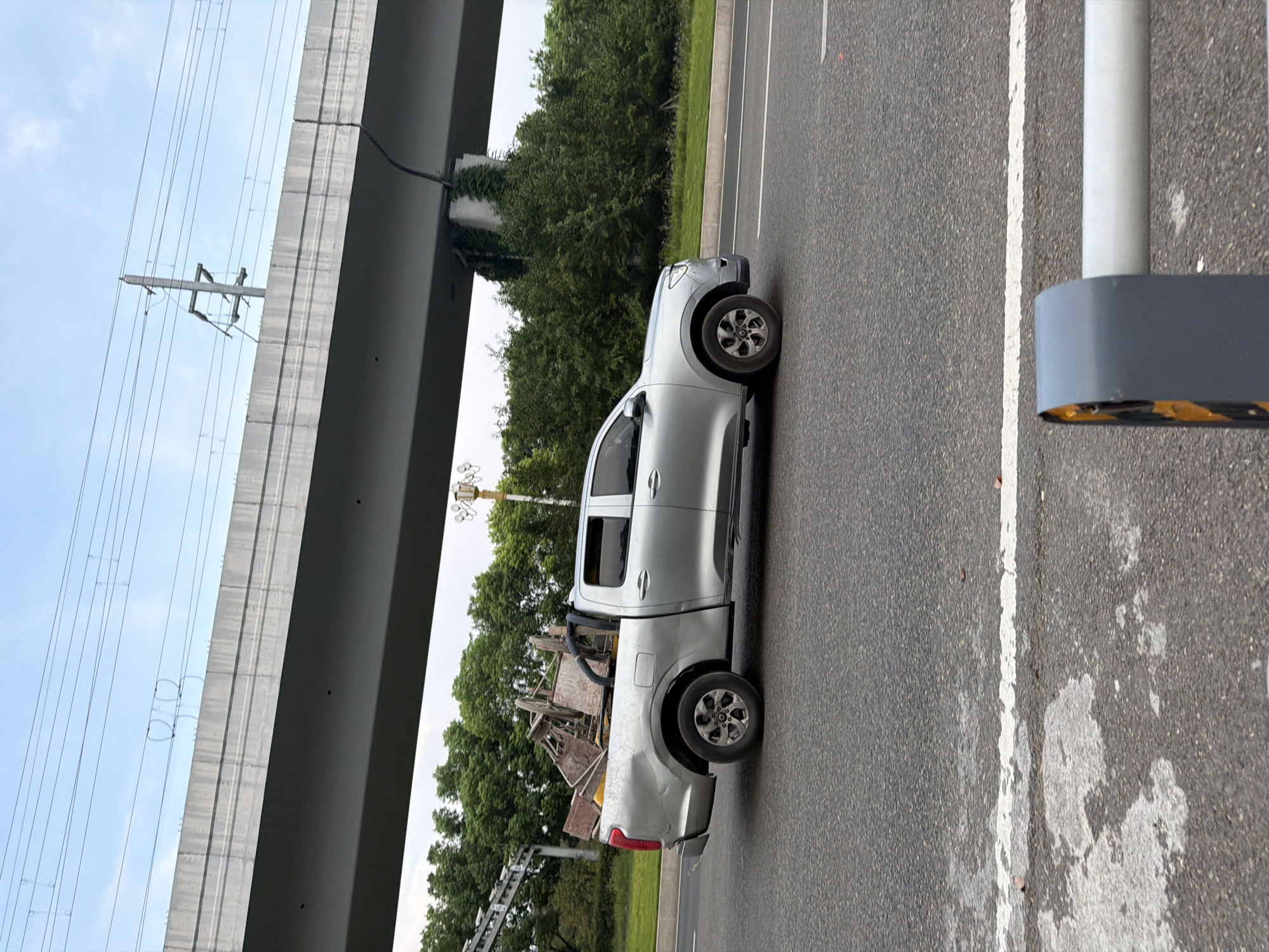 A silver pickup truck is driving on a road in Jiangjin, China, beneath an elevated highway. The truck is loaded with what appears to be dismantled wooden structures and some yellow plastic barrels in its bed. The truck is a double-cab model with a black roll bar behind the cab. The road surface is asphalt with white lane markings. Trees and vegetation line the side of the road, and a concrete support pillar for the elevated highway is visible, partially covered in ivy. The sky is overcast with some visible power lines and insulators strung along the elevated highway. In the foreground, a gray and yellow barrier is partially visible.