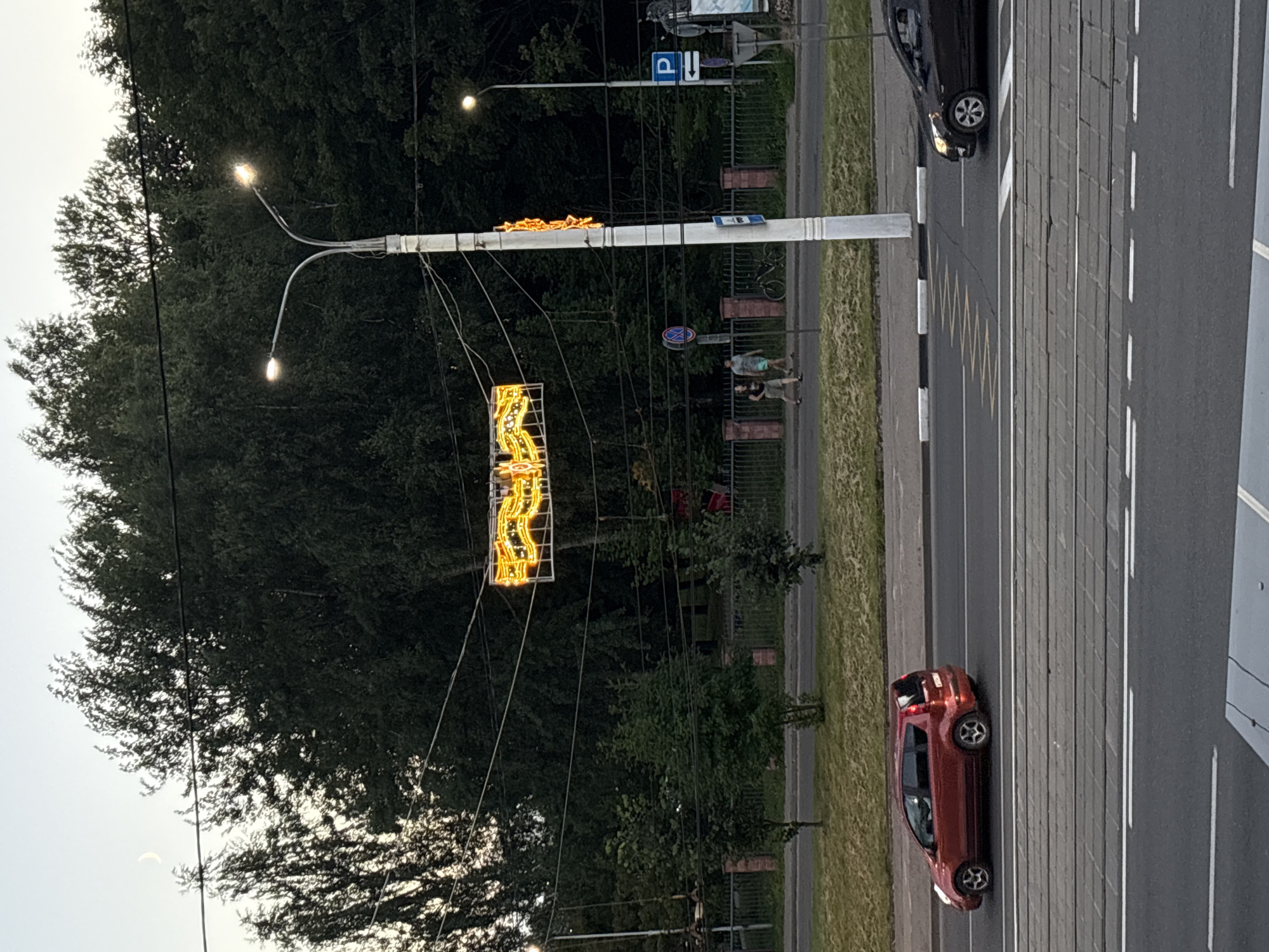 The image depicts a street scene, possibly in Vitsebsk, Belarus, at dusk. A decorative, illuminated sign, possibly a string of lights forming a pattern, is suspended between power lines above a road. A streetlight with two lamps stands to the right of the sign. In the background, dense trees line the street, with a faint crescent moon visible in the sky above them.
On the sidewalk, two individuals are walking. Below them, a red hatchback car is seen driving on the road, and a black car is also visible further down the road. Tram tracks are present in the foreground. A fence separates the sidewalk area from a grassy verge. Signage, including a parking sign, is visible on the right side of the image.