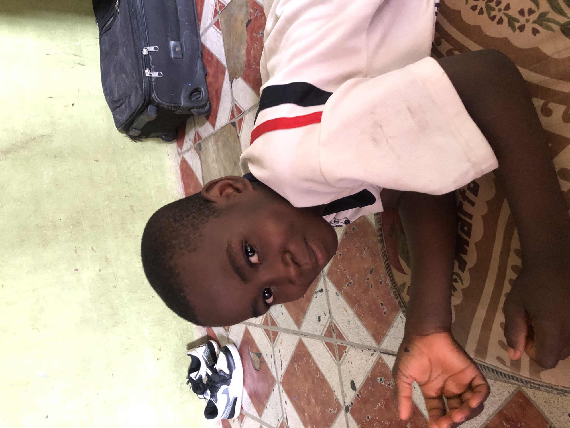 A young Black child, likely a boy, is lying on his back on a tiled floor, looking directly at the camera with a neutral expression. He has short, dark hair and is wearing a white short-sleeved top with black and red stripes on the left sleeve. His left arm is bent, while his right arm is extended, with his hand resting on the floor.
The floor is composed of two main sections: a large area of patterned tiles with alternating reddish-brown and off-white/light brown diamond shapes, and a plain light green or yellowish-green surface to the left. Some of the patterned tiles appear worn or chipped. A dark, patterned mat or rug with a floral design is partially visible under the child's right side, overlapping the tiled floor.
In the upper left portion of the image, a black, wheeled suitcase is partially visible, appearing somewhat dusty or worn. A pair of black and white sneakers is on the floor near the child's feet, in the bottom left. The scene depicts an indoor setting, possibly a home. No visible text is present in the image.
