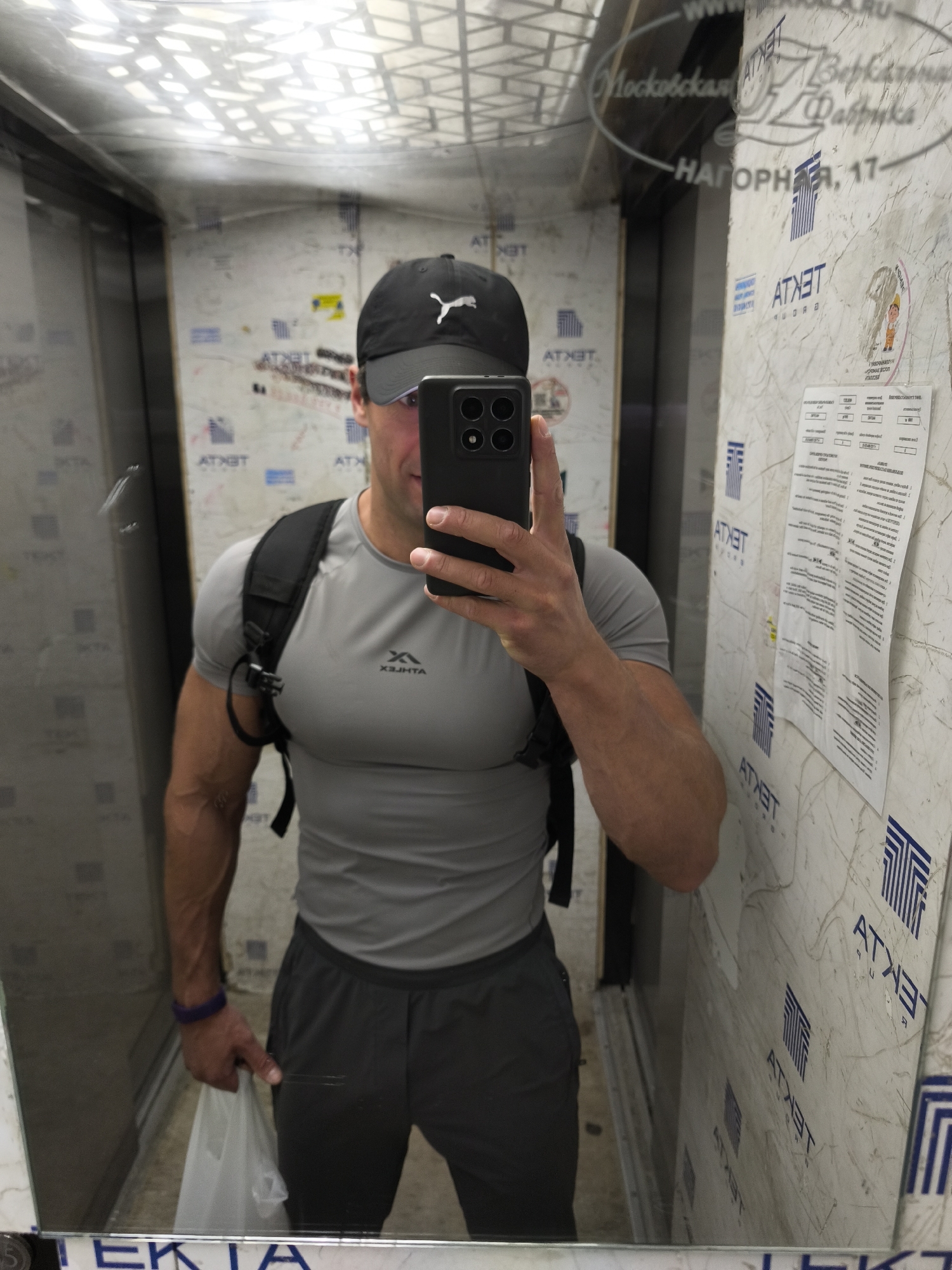 A muscular man in a gray t-shirt, dark shorts, and a black baseball cap takes a mirror selfie in an elevator. He is wearing a black backpack and holding a white plastic bag in his left hand. The elevator walls are covered in white paper with blue and black lettering, appearing to be informational posters or advertisements, some with logos that read "ATXET". Reflections in the mirror show the elevator interior, including a metallic door and a patterned ceiling. Text visible on the walls includes "Московская Фабрика" and "НАГОРНАЯ, 17", suggesting the location is Moscow, Russia. The lighting is artificial and typical of an indoor elevator. The man's expression is neutral.