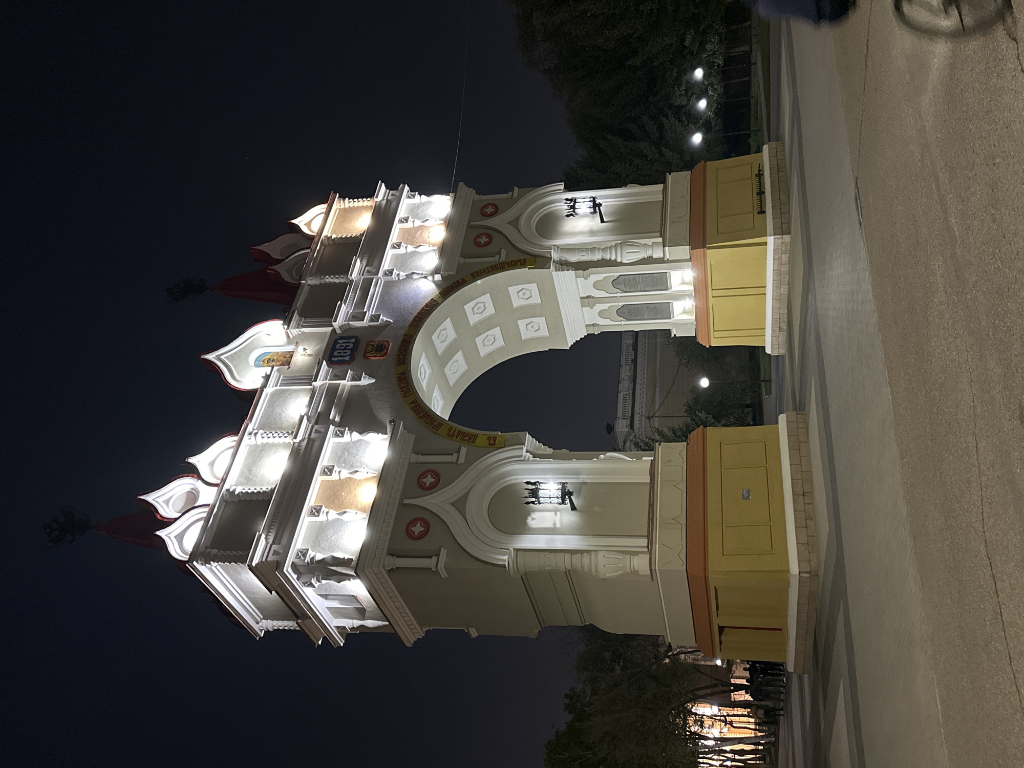 At night, a brightly lit triumphal arch stands prominently in Blagoveshchensk, Russia. The architectural structure, constructed of white stone and yellow panels, features a large arched opening. The archway's upper portion includes multiple levels, with intricate detailing and red, teardrop-shaped accents at the top. Light fixtures illuminate the arch, highlighting its design. Pedestrians are walking on the paved walkway, and trees provide a backdrop to the scene.