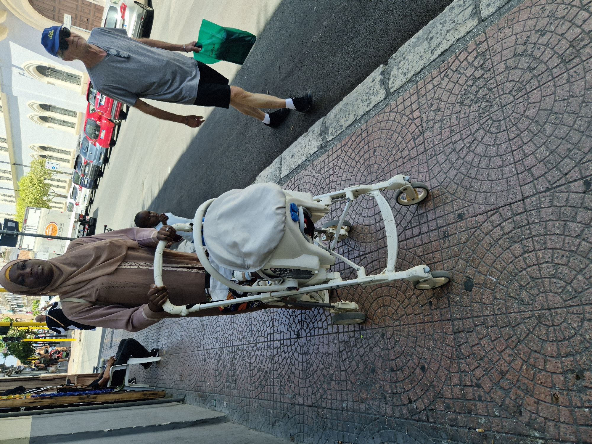 In Taranto, Italy, on a sunlit sidewalk paved with red and grey circular stone patterns, a woman wearing a light brown hijab and dress pushes a white baby stroller. Next to her, a young child peeks over the stroller's edge. Further down the sidewalk, a man wearing a grey t-shirt, black shorts, white socks, and black shoes walks away from the camera, carrying a green shopping bag. He also wears sunglasses and a blue baseball cap. A line of parked cars, including a red Fiat, are visible on the street. The scene takes place in an urban setting, with buildings and streetlights in the background.