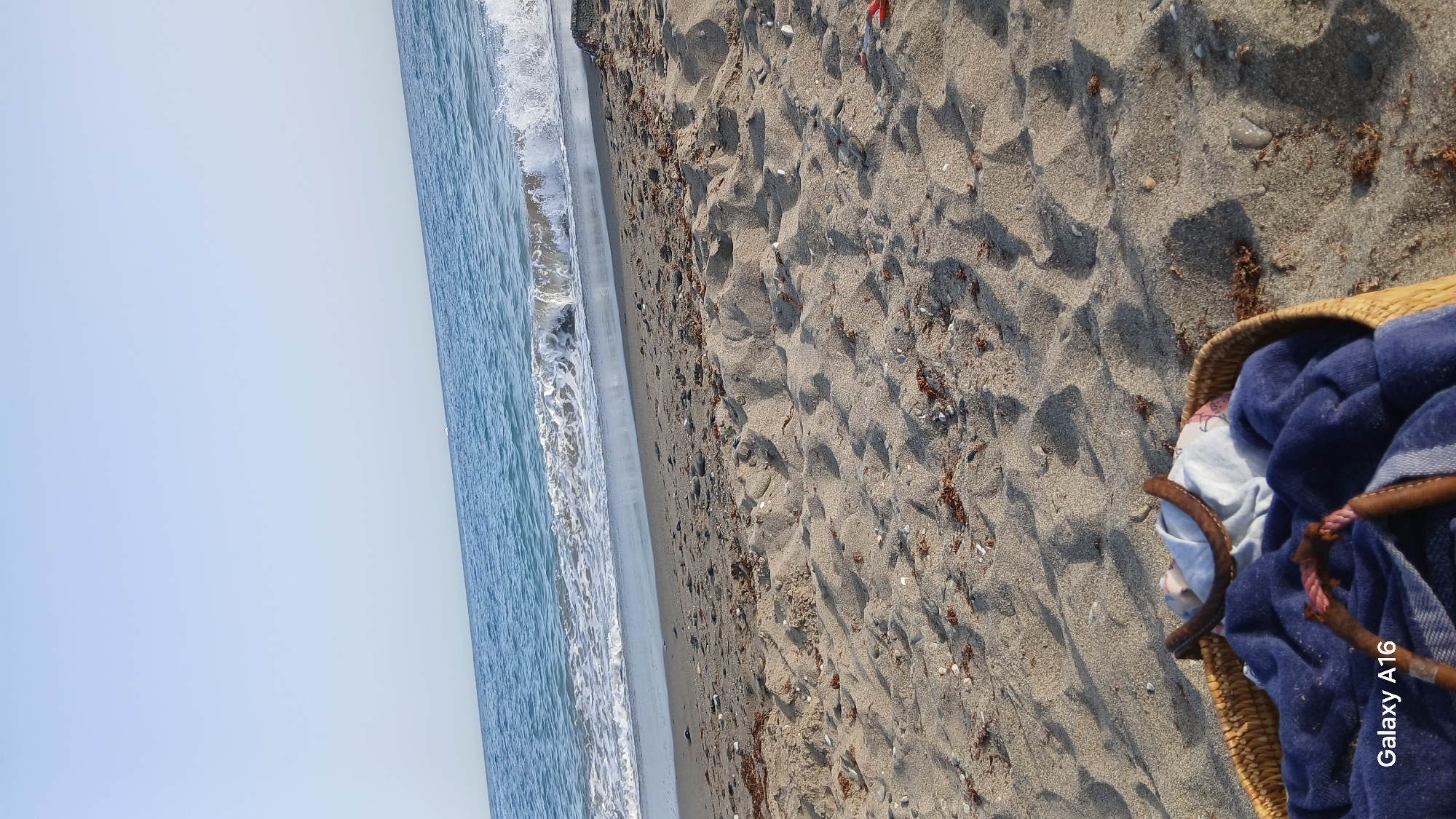 The image depicts a serene beach scene. In the foreground, a woven basket holds a blue towel and some other folded items. The sand is a light brown color, with scattered seaweed and small pebbles. The ocean stretches out in the background, with gentle waves lapping at the shore. The sky is a clear, pale blue. The image is taken from a low angle, looking towards the sea. There are no people visible in the frame. The overall impression is one of peace and relaxation.