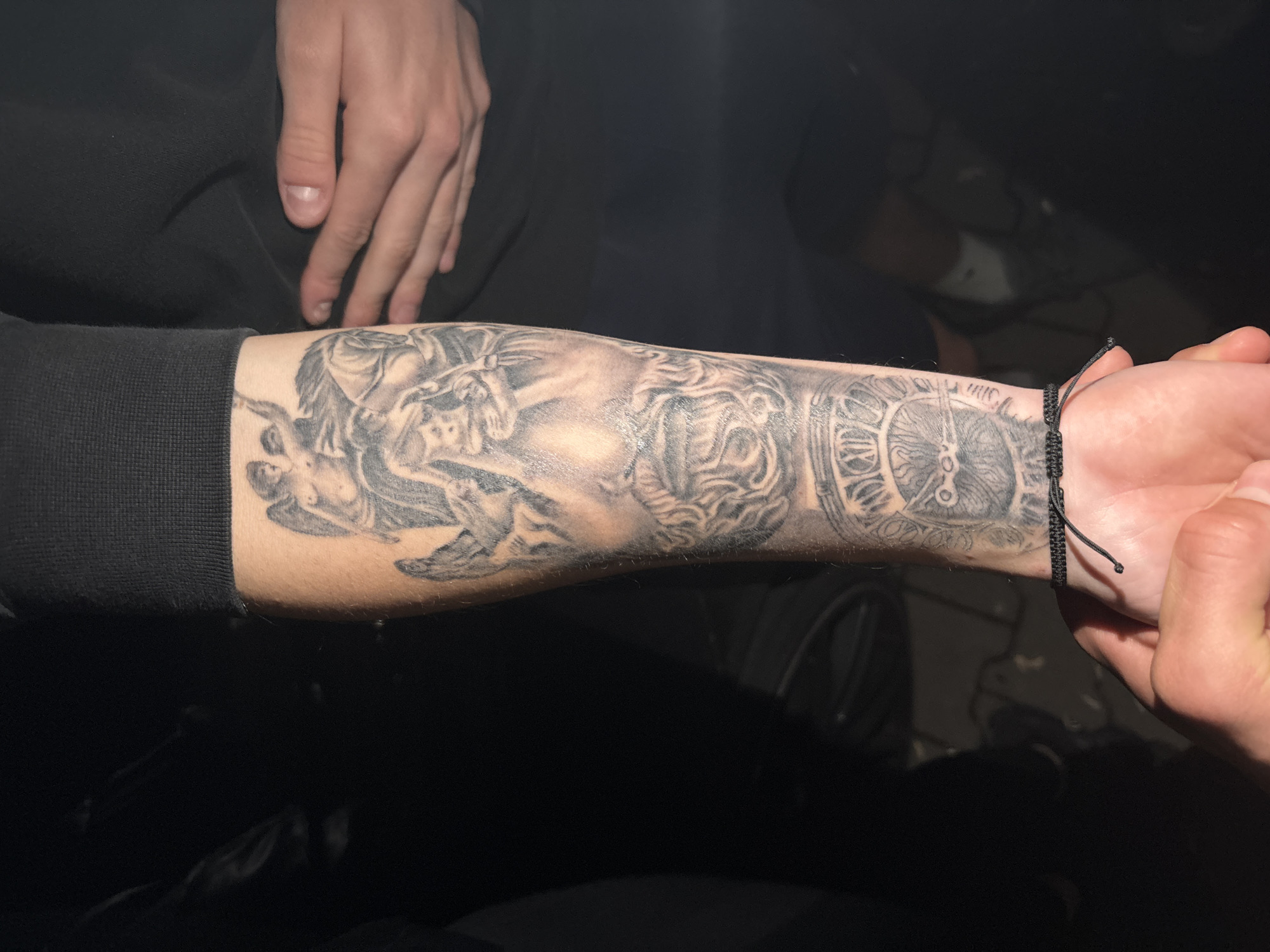 A close-up shot focuses on a person's arm displaying a full tattoo sleeve in grayscale, featuring imagery of angels, and a clock. A black string bracelet is worn around the wrist. The person is wearing a black, long-sleeved shirt or sweatshirt. Another person's hand is partially visible, touching the wrist. The setting appears to be indoors with a dark, possibly dimly lit environment. The photo does not contain any information about the city of Zlatograd, Bulgaria.