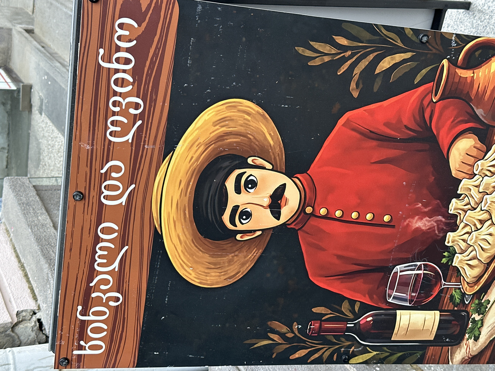 The image shows a close-up of a restaurant sign, likely in the city of Tbilisi, Georgia. The sign features a cartoonish illustration of a man wearing a straw hat and a red shirt with gold buttons. He has black hair and a pronounced mustache. To his right are a clay wine jug, a plate of khinkali (Georgian dumplings), a wine bottle, and a partially filled wine glass. A sprig of green garnish also sits near the wine. The sign's background is black, and stylized gold branches are placed near the jug and wine. The left side of the sign has a wood-grain effect, with white Georgian lettering vertically arranged. The sign is attached to a wall or post, with what appears to be gray concrete stairs visible to the left. The image is slightly tilted.