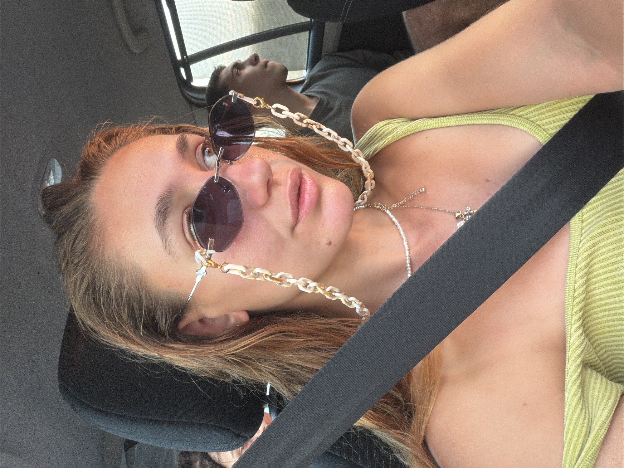 A young woman, facing the camera, sits in the passenger seat of a vehicle. She wears sunglasses with a pearl and chain strap, a pearl necklace with a pendant, and a light green ribbed tank top. Her long, reddish-brown hair is loose. A black seatbelt is diagonally across her chest. In the background, to her right, a young man sits in the back seat, looking out the window. The interior of the car, including the seats and ceiling, is visible. The lighting suggests it is daytime, possibly late afternoon given the warm glow. The context of "Παλαιοκώμη, Greece" indicates the geographical setting, implying the car is on a road in or near this location.