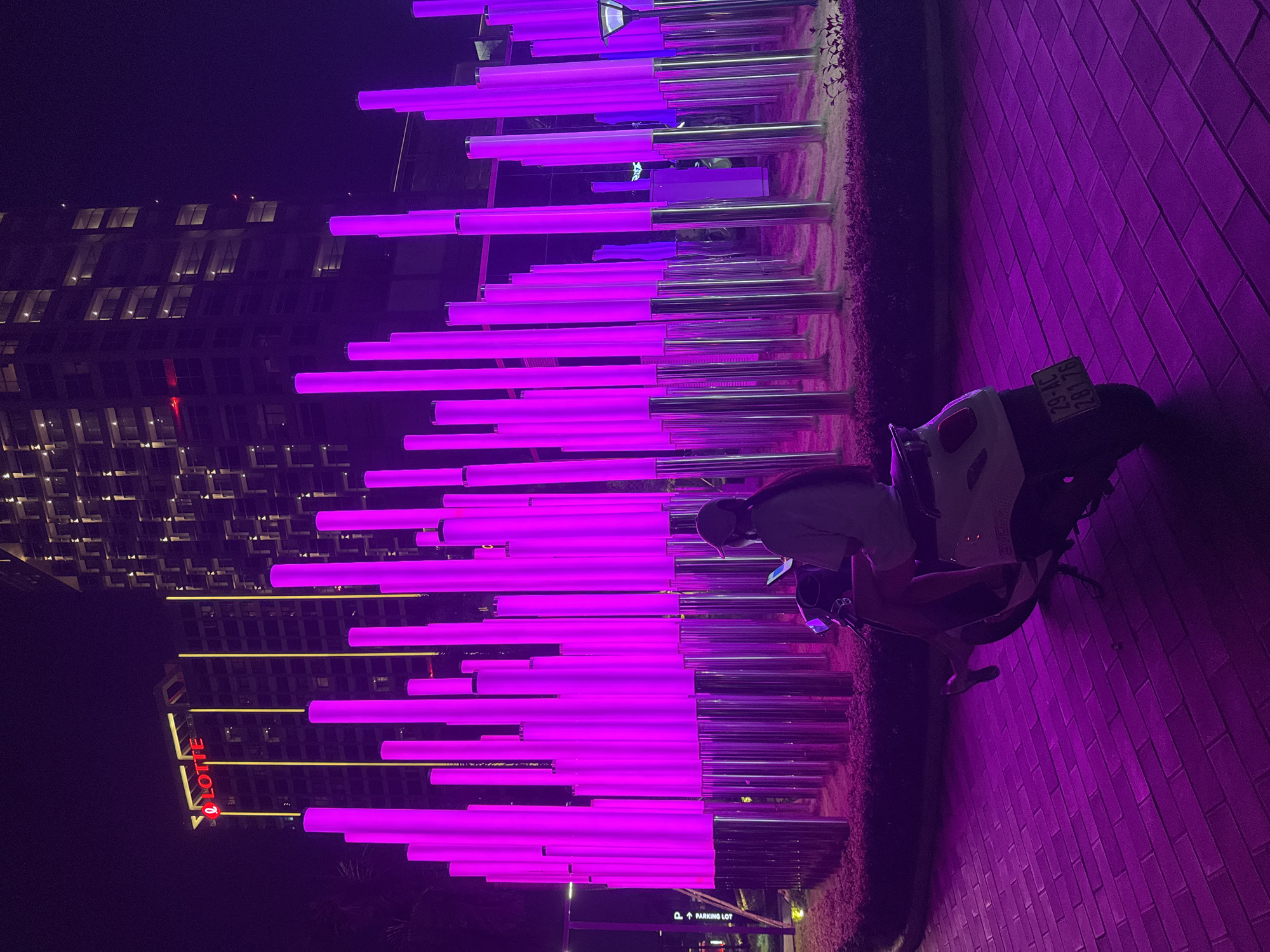 The image captures a nighttime scene in Hanoi, Vietnam, illuminated by vibrant purple lights. The foreground features a modern sculpture composed of numerous vertical, cylindrical elements bathed in a bright pink-purple glow. Interspersed with the illuminated tubes are reflective silver cylinders. To the right of the sculpture, a motorcycle is parked on a brick-paved walkway, also bathed in the ambient purple light. A person wearing a cap is seated on the motorcycle, looking down at a mobile device.
In the background, a tall building with many illuminated windows stands against the dark sky, suggesting an urban setting. The building's architecture appears modern and multifaceted. A bright red and yellow "LOTTE" logo is visible on the building, providing a specific location cue. The overall impression is one of a lively, contemporary urban environment at night, enhanced by decorative lighting. The time of day is clearly night due to the dark sky and artificial lighting. The weather appears clear, as there are no visible signs of rain or fog. The scene evokes a sense of urban nightlife and technology. Visible text includes "LOTTE" on the building and a license plate on the motorcycle, which reads "29-AC 281.77". A directional sign with an arrow and text "PARKING LOT" is also visible in the lower portion of the image.