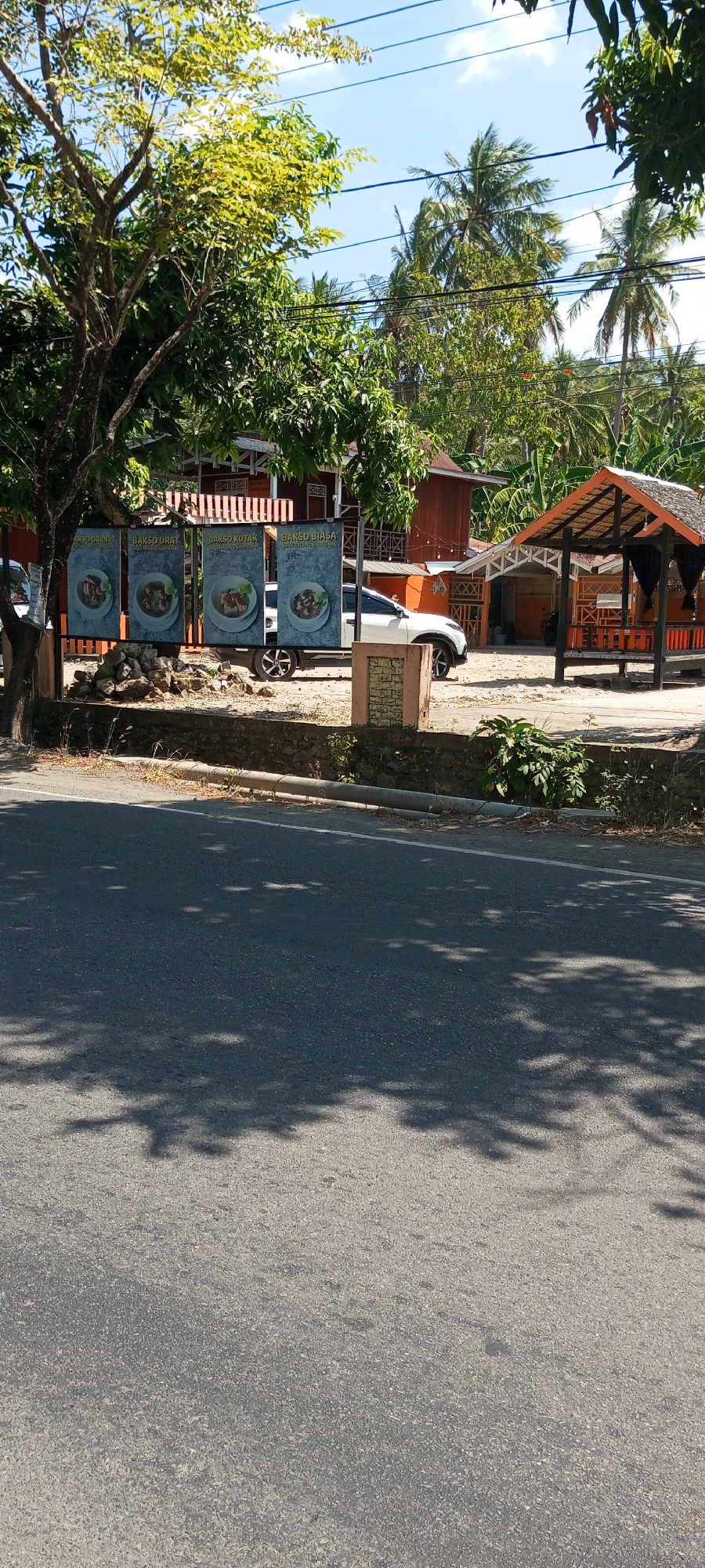 This image captures a scene in Lempa, Indonesia, featuring a roadside establishment with several signs displaying food items, specifically "BAKSO GHANAT," "BAKSO URAT," "BAKSO KOTAK," and "BAKSO BIASA," each accompanied by a picture of a bowl of food. A white SUV is parked behind the signs. In the background, there is a building with a reddish-brown exterior and a thatched-roof gazebo. The environment is tropical, with lush green trees and palm trees visible, along with a clear blue sky. The foreground shows a paved road with tree shadows cast across it. The overall impression is of a casual eatery situated in a semi-rural, tropical setting.