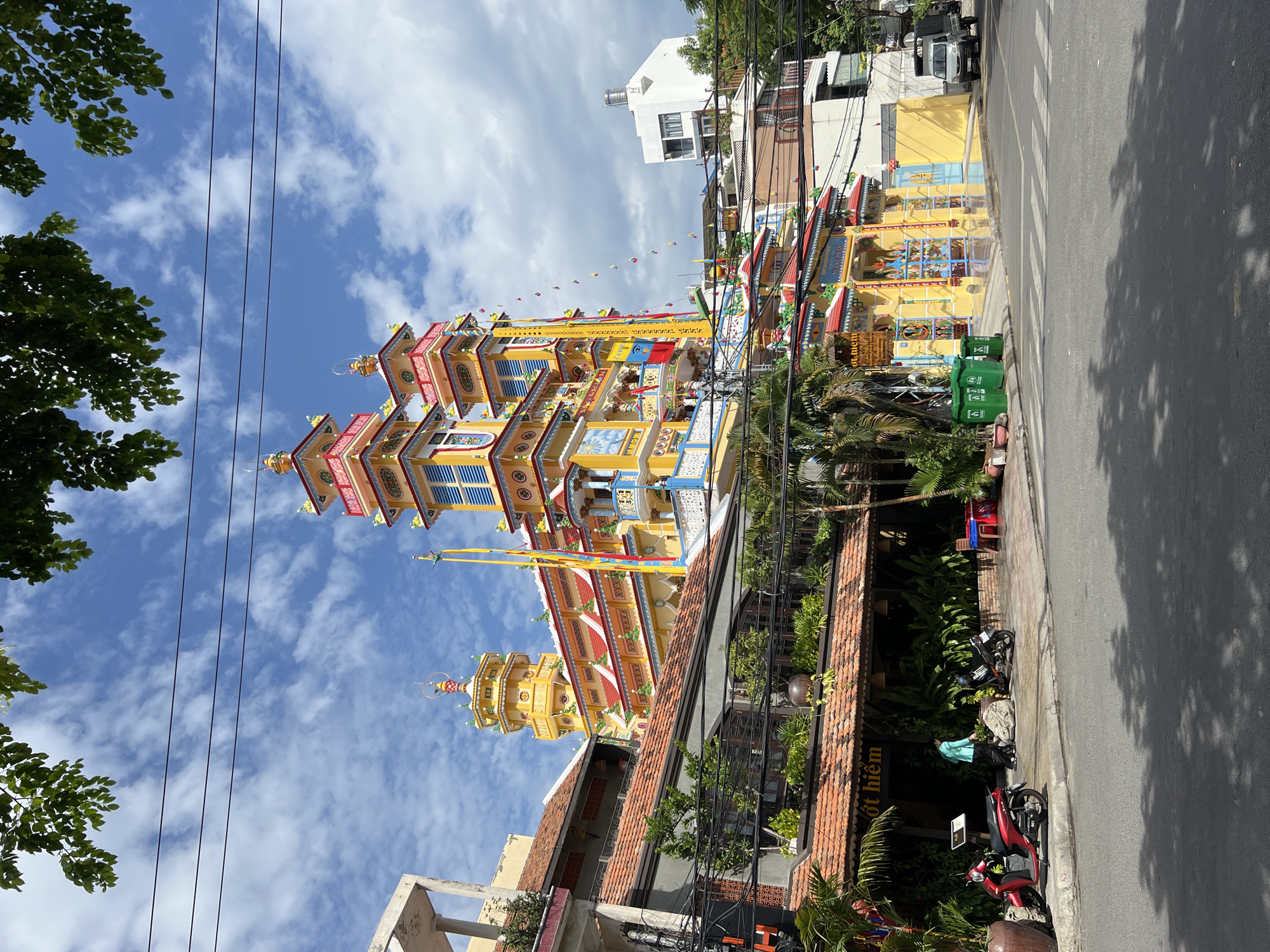 This image captures a vibrant scene in Nha Trang, Vietnam, focusing on a highly ornate Buddhist pagoda situated on a city street. The pagoda's architecture is a striking display of color, with prominent yellow, red, and blue hues dominating its multi-tiered structure. Multiple spires and decorative elements adorn the building, some topped with golden finials. Small flags and banners, many in yellow and red, are visible, adding to the festive atmosphere.
In the foreground, a street with a pedestrian crossing is visible, with shadows from overhead trees cast upon the asphalt. To the left, a red scooter is parked near a building with a reddish-brown tiled roof. A person in a turquoise top is walking away from the scooter towards a darker motorcycle parked on the sidewalk. Beside the parked motorcycles, lush green plants are visible, suggesting a verdant environment within the urban setting. Several green trash receptacles are placed near the entrance of another building to the right of the pagoda. Further to the right, a white truck is parked on the street. The sky above is a mix of blue with scattered white clouds. Utility wires are strung across the upper portion of the image. The buildings surrounding the pagoda, including the one with the tiled roof, appear to be a mix of residential and commercial establishments, indicative of a bustling city neighborhood.