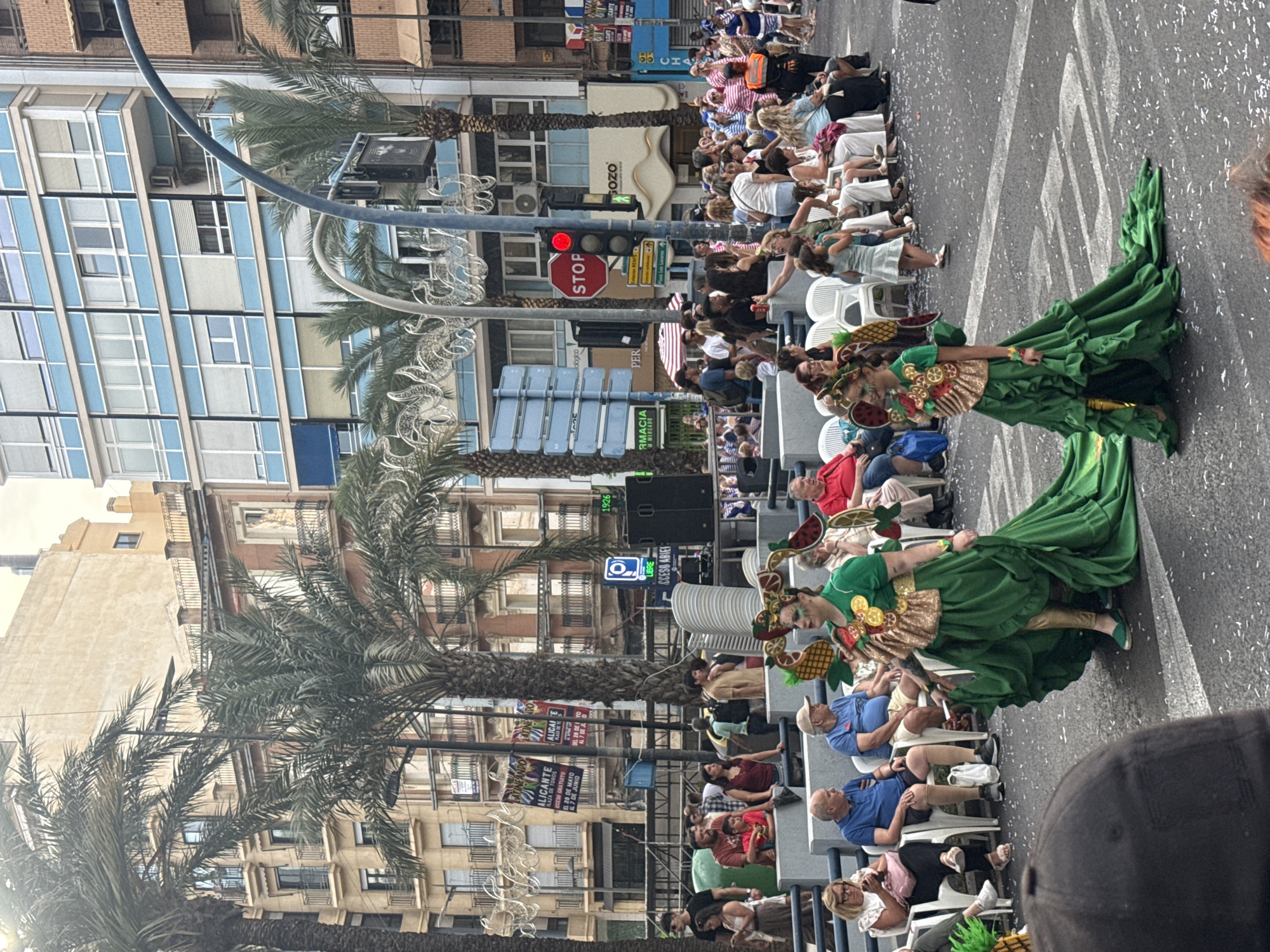 The image depicts an outdoor urban scene during a daytime event, likely a parade or festival, in Alicante, Spain. Two individuals in elaborate green and gold costumes with fruit-themed headpieces are walking down an asphalt street scattered with white confetti. The leading figure wears a long, ruffled green gown, and a smaller figure behind them wears a similar, but shorter, costume with a long train. Spectators, a mix of adults and children, are seated on white plastic chairs or standing behind barriers on both sides of the street, observing the procession.
In the background, a mix of architectural styles is visible: older, multi-story buildings with ornate balconies and a beige facade, alongside a modern building with distinctive blue and white horizontal stripes and numerous windows. Palm trees line the street. Street infrastructure includes a traffic light displaying red, various street signs (one indicating "LIBRE" and "ACCESO ARBIEL"), and decorative white string lights overhead. Banners advertising "ALICANTE" and dates "29 DE MAYO AL 7 DE JUNIO" are prominently displayed, further indicating the location and context of the event. A large speaker is positioned on a raised platform on the right side. The bottom left foreground is partially obscured by a dark cap.